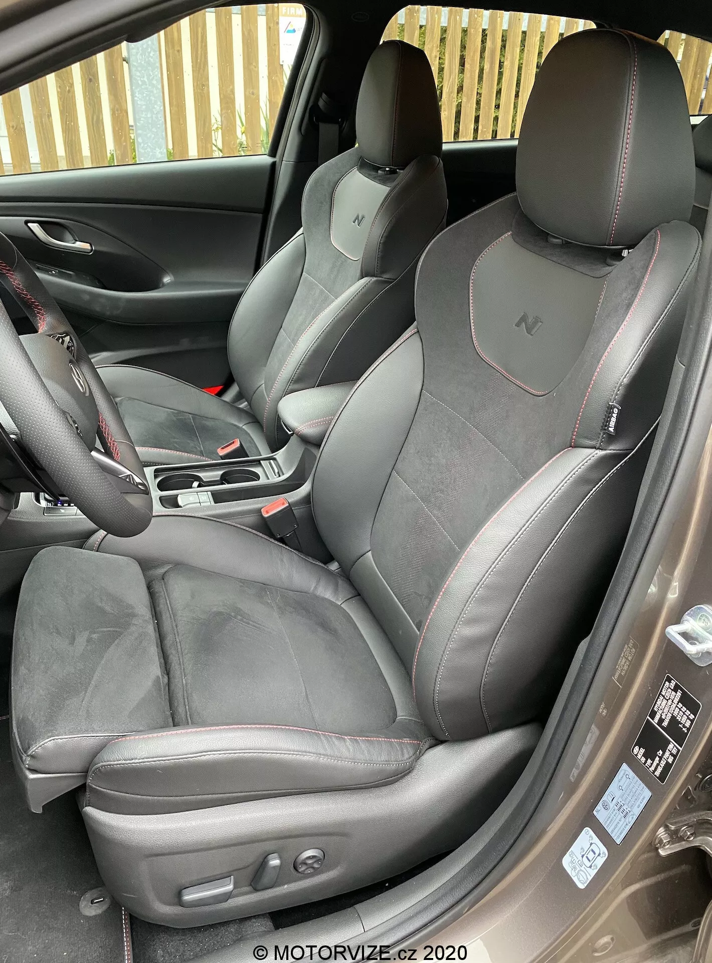 Esta imagen detallada muestra el interior de la cabina delantera de un Hyundai i30 2020 (i30-kombi) desde un ángulo ligeramente elevado fuera de la puerta delantera del pasajero. La puerta del pasajero abierta revela el lado de los asientos deportivos premium tapizados en gamuza o alcantara y cuero con costuras contrastantes. Los reposacabezas integrados con el logotipo 'N' insinúan el nivel de equipamiento del automóvil. El asiento del conductor presenta botones de control eléctrico para ajustes de asiento con botones de control eléctrico para ajustes de asiento con botón de control. Unidos para una mejor vista general de las funciones tecnológicas y de confort del automóvil.