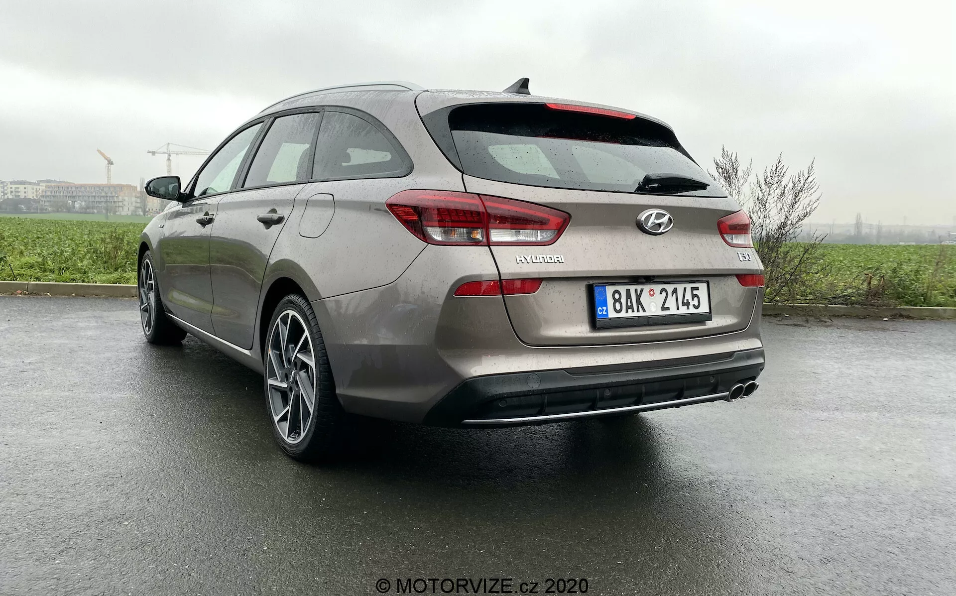 Vista tres cuartos trasera del Hyundai i30 Kombi 2020 desde un ángulo bajo que muestra detalles del diseño, incluyendo luces traseras, paragolpes trasero, portón trasero, cámara de estacionamiento trasera, llantas de aleación y reflectores en una superficie mojada por la reciente lluvia, destacando los sistemas avanzados de asistencia al conductor y el atractivo estético del vehículo.