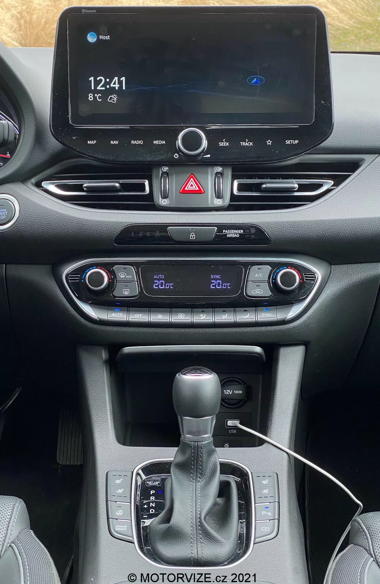 Vista frontal de la consola central del Hyundai i30 Kombi 2020 desde los asientos frontales, mostrando el sistema de información y entretenimiento digital que indica la hora (12:41), la temperatura (8°C) y el icono de Bluetooth. Debajo, se encuentra el botón de advertencia de peligro, el control de clima de doble zona con ajustes de temperatura separados (conductor 22°C, pasajero 20°C), botón de arranque y parada del vehículo, palanca de cambio automática con modo manual, posibles portavasos/espacio de almacenamiento, puerto de conectividad con cable visible y diseño de materiales mixtos del tablero y la consola. Marca de agua de derechos de autor '@ MOTORVIZE.cz 2021' en la parte inferior.