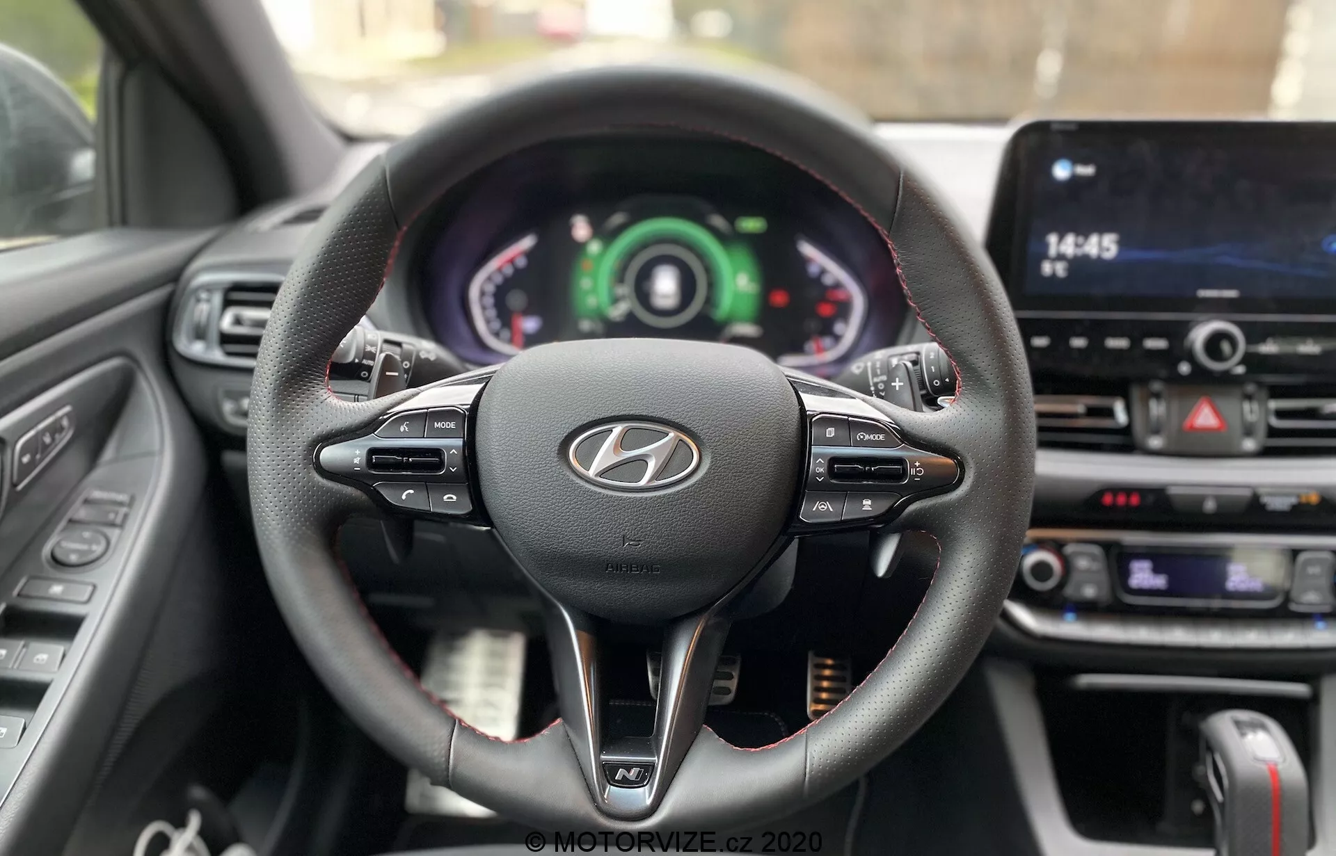 Vista desde la perspectiva del conductor del volante y el tablero del Hyundai i30 2020, que muestra un diseño de tres radios envueltos en cuero con controles multifunción, un logotipo prominente de Hyundai y un clúster de instrumentos con pantallas tanto analógicas como digitales. La pantalla táctil de infoentretenimiento central y los botones de control del clima destacan la tecnología avanzada en el coche y los elementos interactivos del conductor.