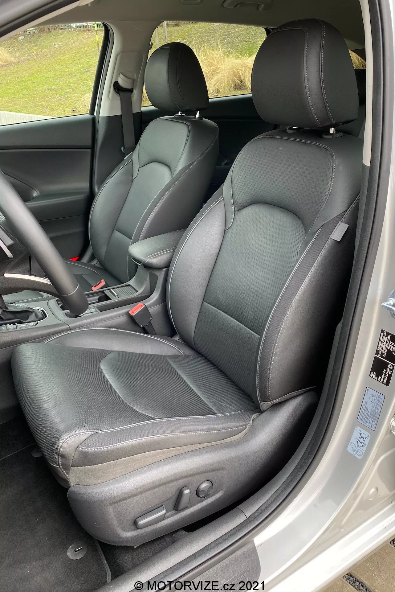 Una vista detallada del interior frontal de un Hyundai i30 Kombi 2020, centrada en el lado del conductor. La perspectiva es desde ligeramente por encima del nivel del asiento, mirando en diagonal hacia abajo dentro de la cabina como si se entrara desde la puerta trasera. El asiento del conductor de cuero negro se destaca con costuras visibles y controles de ajuste electrónico en el lado. El cinturón de seguridad y el tercio izquierdo del volante de tres radios son visibles. Son visibles parcialmente partes de la consola central, incluida la palanca de cambios automática en posición 'park', así como parte del tablero y una ventilación. La cabina tiene un esquema de color negro monotono, con texturas variadas en los asientos, los paneles de las puertas y el tablero. La foto está bien iluminada con luz natural, lo que sugiere un ambiente nublado o sombreado.