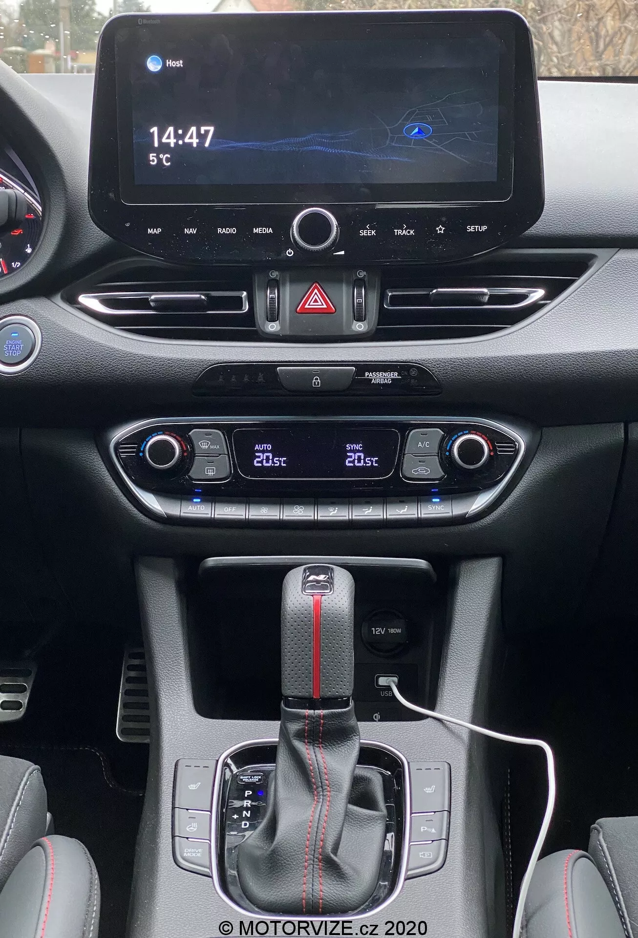 Perspectiva del conductor del Hyundai i30 Kombi 2020 en la consola central. En la parte superior, una pantalla de infoentretenimiento moderna muestra un mapa de navegación, un reloj digital que marca las '14:47' y una temperatura ambiental de '5°C'. Debajo están el botón de luces de emergencia y las rejillas de ventilación. Más abajo, el panel de control del clima de doble zona muestra '20°C' para ambos lados. La palanca de cambios automática con funda de cuero y una franja roja indica un acabado deportivo. Un cable de carga blanco está conectado cerca de la palanca de cambios, rodeado de controles probablemente para modos de conducción o control de tracción. Los materiales combinan superficies lisas y texturizadas con costuras contrastantes, enfatizando un diseño interior contemporáneo y deportivo.