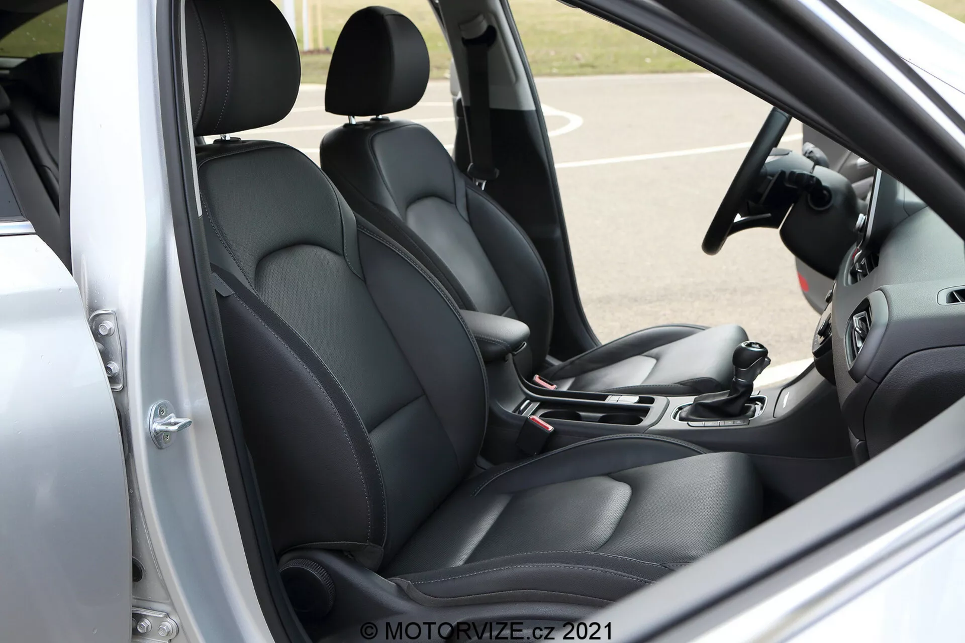 Vista de ángulo lateral cercano del interior de la cabina delantera del Hyundai i30 Kombi 2020. La imagen muestra el asiento del conductor y parte del asiento del pasajero, ambos tapizados en negro con un diseño contorneado para mayor comodidad y soporte. Visible desde el lado del conductor con la puerta abierta, la escena incluye una palanca de cambios, freno de mano manual y parte de los sistemas de infoentretenimiento y control de clima, insinuando características tecnológicas modernas. El panel de la puerta del conductor, que muestra los controles de las ventanas eléctricas y posiblemente los ajustes del espejo, agrega al diseño funcional general. El volante, aunque parcialmente cortado, insinúa controles integrados para una mejor interacción vehicular. Marca de agua con 'MOTORVIZE.CZ 2021' en la esquina inferior derecha.