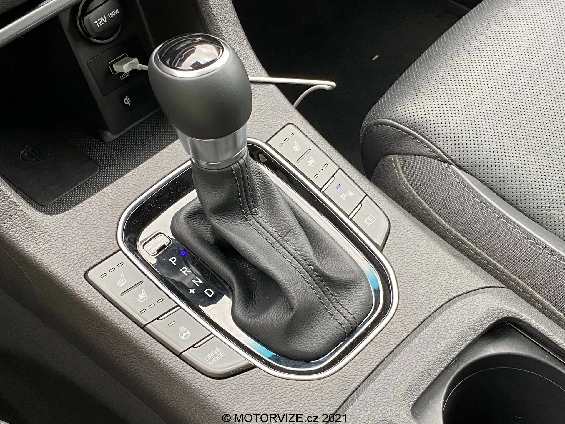 Detalle en primer plano del cambio automático del Hyundai i30 2020, tomado desde un ángulo de 45 grados. La palanca de cambios está rodeada por un embellecedor negro brillante con una funda de cuero y costuras visibles. El esquema PRND es claramente visible junto con las indicaciones de modo manual o deportivo. Los botones de la izquierda controlan los modos de conducción, mientras que los de la derecha gestionan la estabilidad o el control de tracción del vehículo. También se nota el freno de estacionamiento de operación electrónica. La imagen bien iluminada resalta la calidad premium y el meticuloso diseño de la consola central.