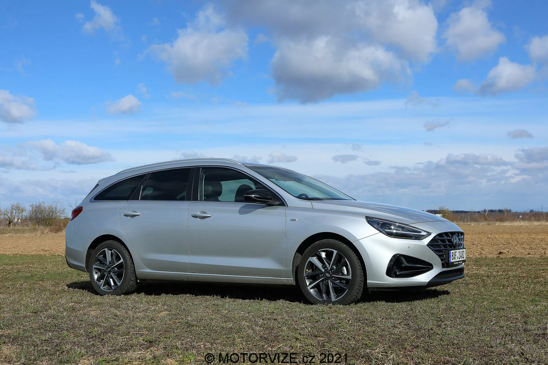 Hyundai i30 2020 Kombi - Coche hatchback moderno en un campo de hierba con un cielo despejado de fondo, ángulo de perfil lateral que resalta la parrilla delantera, el diseño de las ruedas, los faros, los espejos laterales, las ventanas, las manijas de las puertas y la línea del techo; notable por su pintura metálica elegante, ruedas de aleación y características avanzadas de iluminación y aerodinámica.