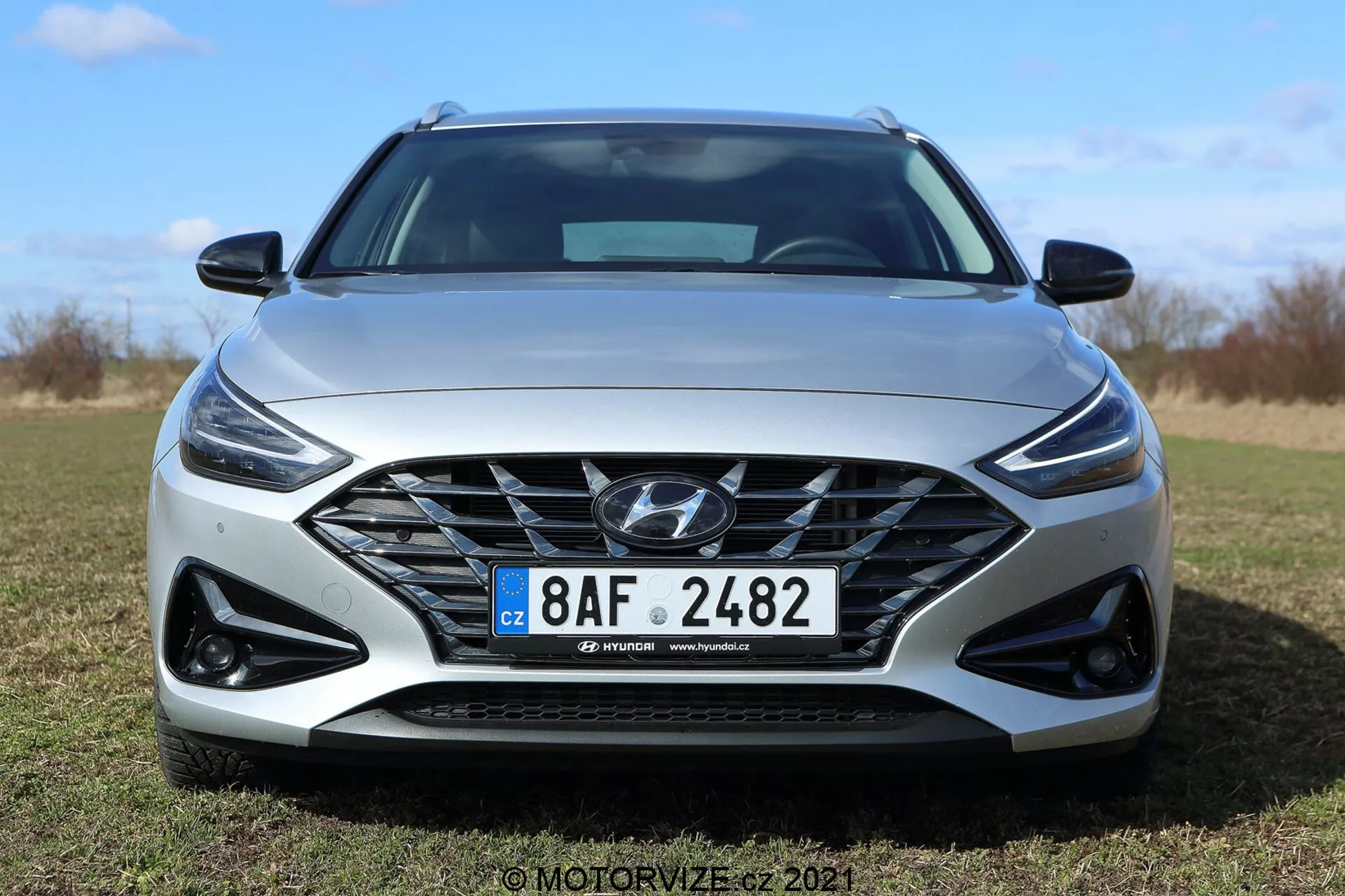 Vista frontal de un Hyundai i30 Kombi 2020 en un día soleado en un entorno al aire libre. El diseño moderno del coche presenta una gran parrilla con una malla intrincada y el logo de Hyundai, faros LED elegantes con luces diurnas y carcasas de luces antiniebla deportivas en negro. El parachoques delantero dinámico y el capó esculpido enfatizan una estética contemporánea y aerodinámica. Son visibles la matrícula de estilo europeo y las posibles llantas de aleación. El color gris claro del coche resalta su estilo sofisticado.