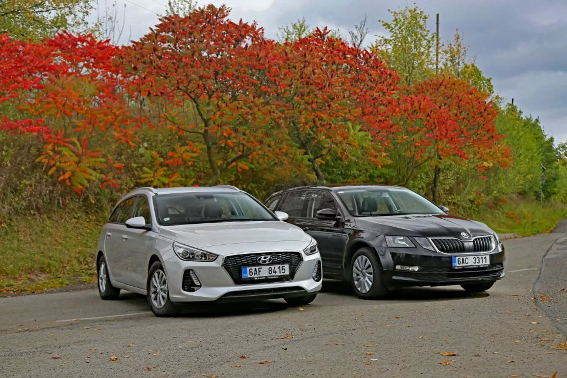 Hyundai i30 hatchback blanco de 2017 y sedán de color oscuro uno al lado del otro en carretera tranquila rodeada de vegetación otoñal, mostrando líneas de diseño y tecnología de iluminación moderna