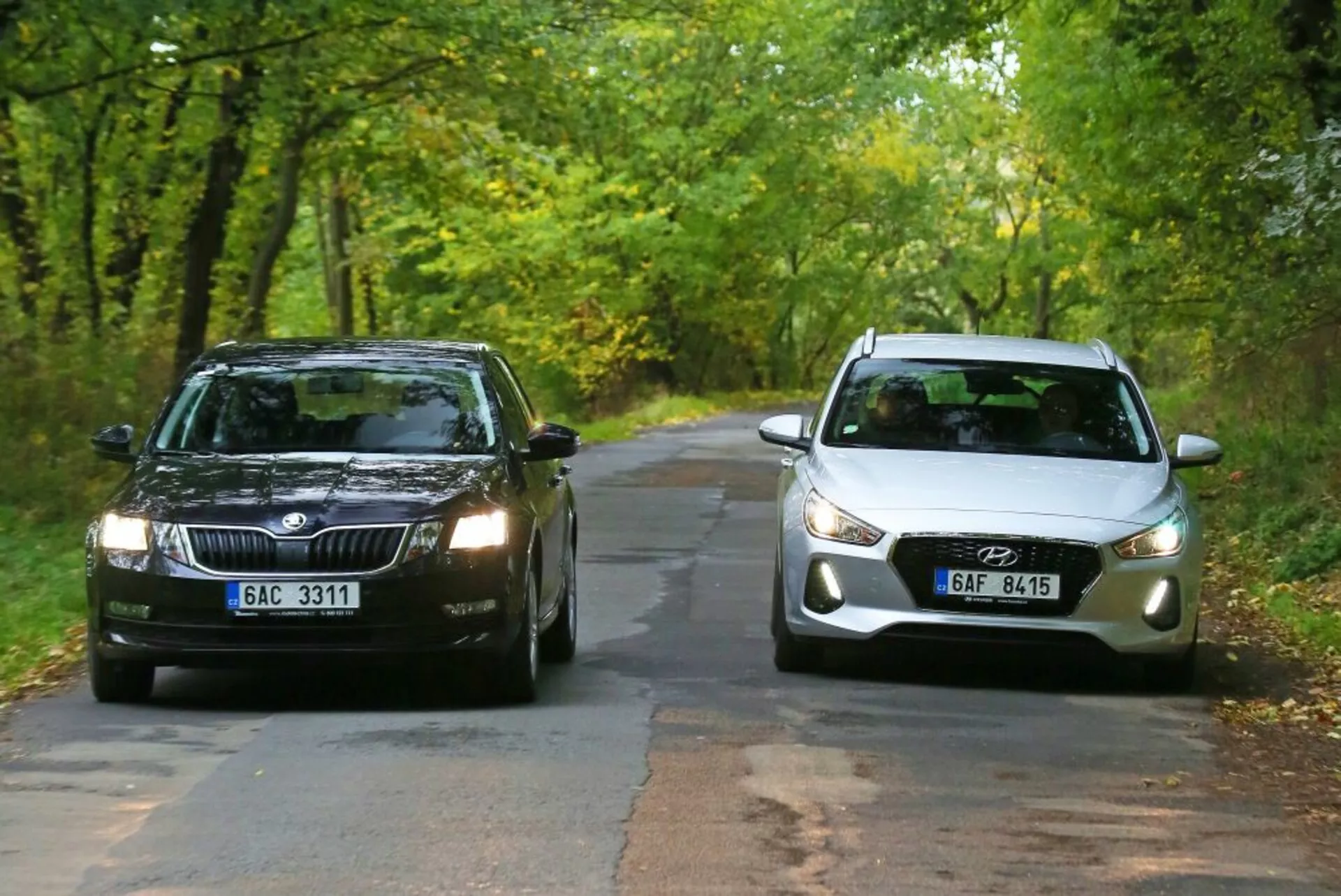 Dos coches enfrentados en un camino rural rodeado de árboles en el inicio del otoño: un SKoda de color oscuro con parrilla rectangular y faros estándar a la izquierda, y un Hyundai i30-N de color claro con frente curvilíneo, faros proyectores modernos y luces de niebla integradas a la derecha, ilustrando una comparación de elementos de diseño de coches tradicionales versus modernos.