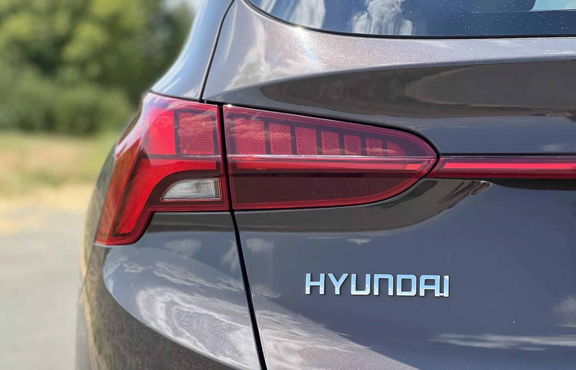 Un primer plano del cuarto trasero del lado del conductor de un Hyundai Santa-Fe 2020, enfocado en el diseño LED en forma de T de la luz trasera y en el distintivo 'HYUNDAI' destacado. El acabado metálico refleja la luz natural del día, resaltando las curvas y líneas de diseño del coche en una toma ajustada y detallada con poca profundidad de campo.