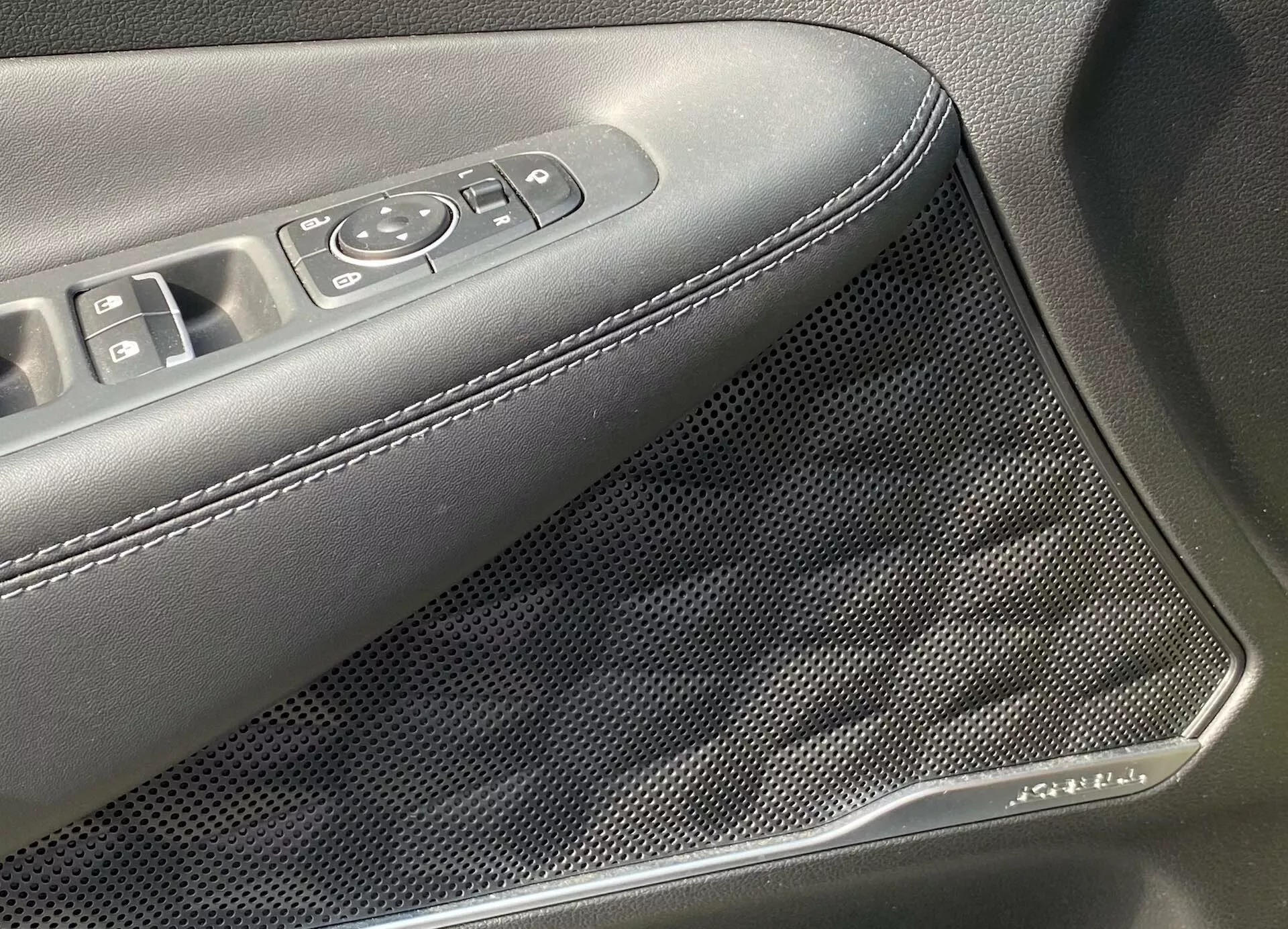 Vista detallada del panel interior de la puerta de un Hyundai Santa Fe 2020, destacando el área con controles de ventanas y espejos eléctricos, así como una rejilla de altavoz con patrón de panal. El reposabrazos está tapizado en material oscuro con costuras blancas. Un adorno similar al cromo adorna la rejilla del altavoz, indicando características avanzadas de audio y de diseño estético.