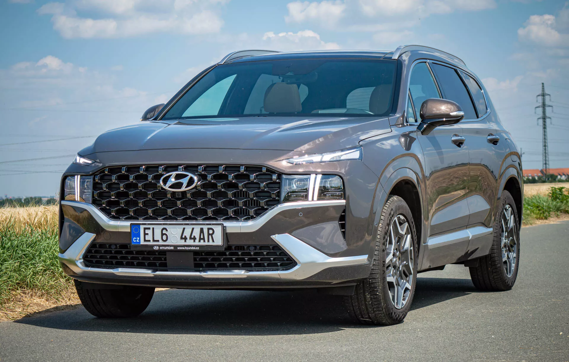 La imagen muestra un SUV Hyundai Santa Fe 2020 estacionado en una carretera de asfalto, capturado desde un ángulo de tres cuartos frontal izquierdo. Se destaca la gran parrilla con un patrón específico de la marca y el logotipo central, los elegantes faros LED y el capó esculpido. El parabrisas es amplio y ofrece una visibilidad suficiente, y el perfil lateral revela llantas de aleación multirradio y el espejo lateral del conductor con señales de giro integradas potencialmente. El vehículo de color oscuro transmite una apariencia sofisticada con sus elementos de diseño modernos, incluidos posibles sensores o cámaras frontales.