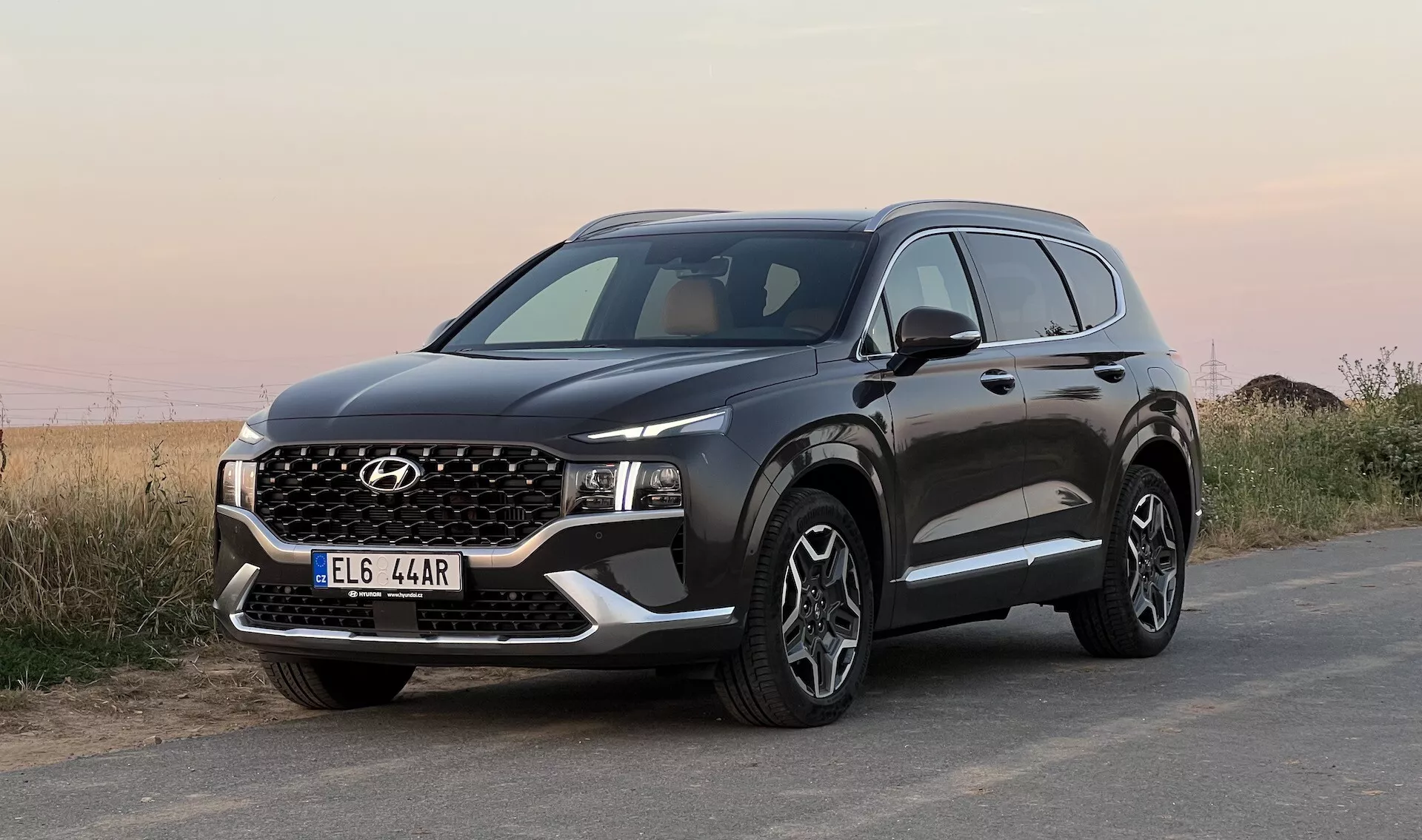 Imagen de un SUV Hyundai Santa Fe 2020 estacionado sobre una superficie de asfalto, tomada desde un ángulo bajo con vista frontal de tres cuartos, mostrando la parrilla delantera, faros LED estrechos, parachoques con luces antiniebla y placa protectora falsa, capó con pliegues angulares, grandes ruedas de aleación de varios radios, protección negra en los arcos de las ruedas y la parte inferior de las puertas, espejo lateral con señales de giro integradas y ventanas con moldura cromada. El fondo presenta un paisaje natural indistinto bajo un cielo abierto.
