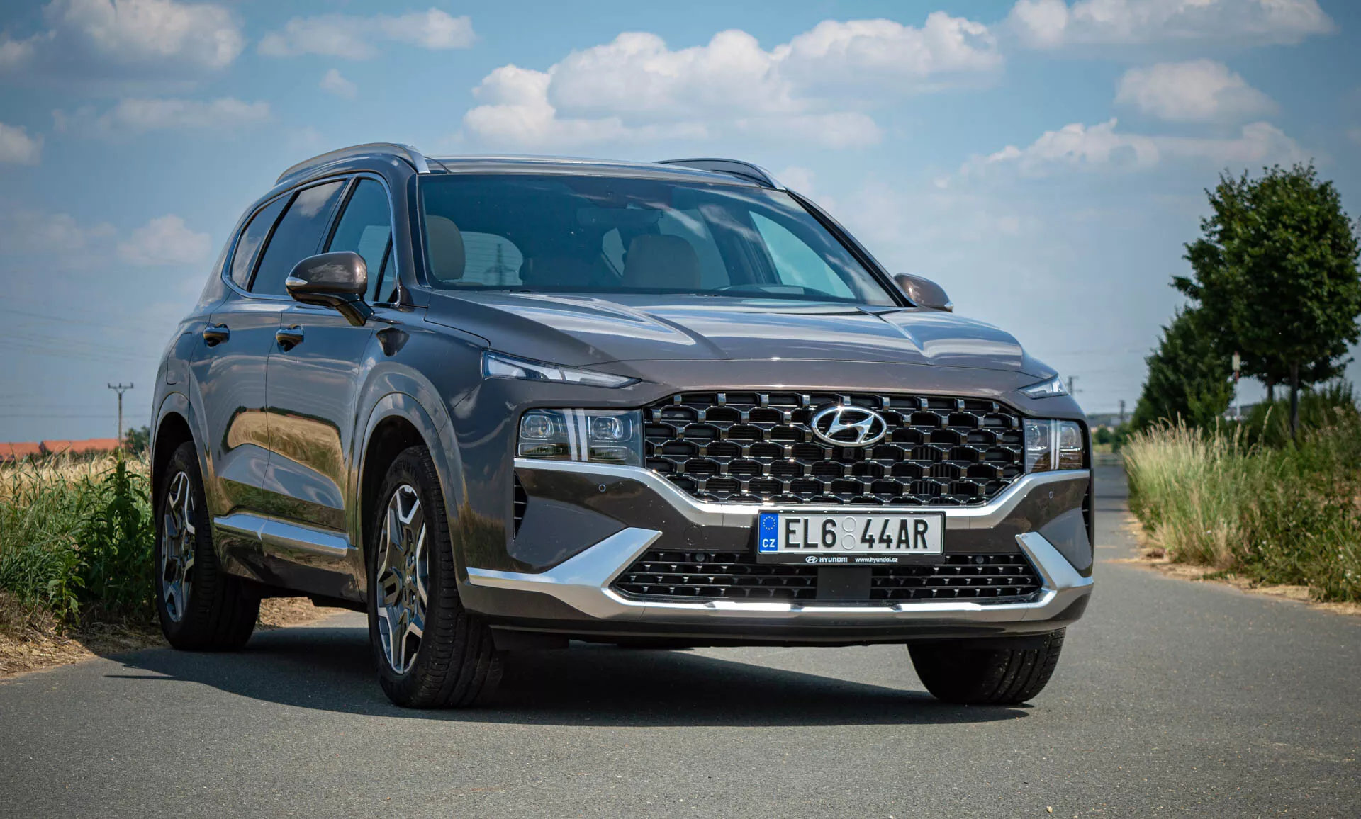 Imagen de un SUV Hyundai Santa-Fe 2020 estacionado en una carretera pavimentada bajo un cielo despejado con vegetación en el fondo. La foto está tomada desde una perspectiva de tres cuartos delanteros, mostrando la fascia frontal del vehículo y el perfil lateral izquierdo. El SUV presenta una parrilla grande de patrón entrelazado con el emblema de Hyundai, faros angulares elegantes LED o HID, llantas de aleación de múltiples radios, un carrocería moderna y aerodinámica con líneas suaves, espejos laterales con señales de giro integradas, ventanas tintadas, color gris oscuro, una placa delantera y parece estar en perfecto estado, lo que sugiere que es nuevo o está bien mantenido.