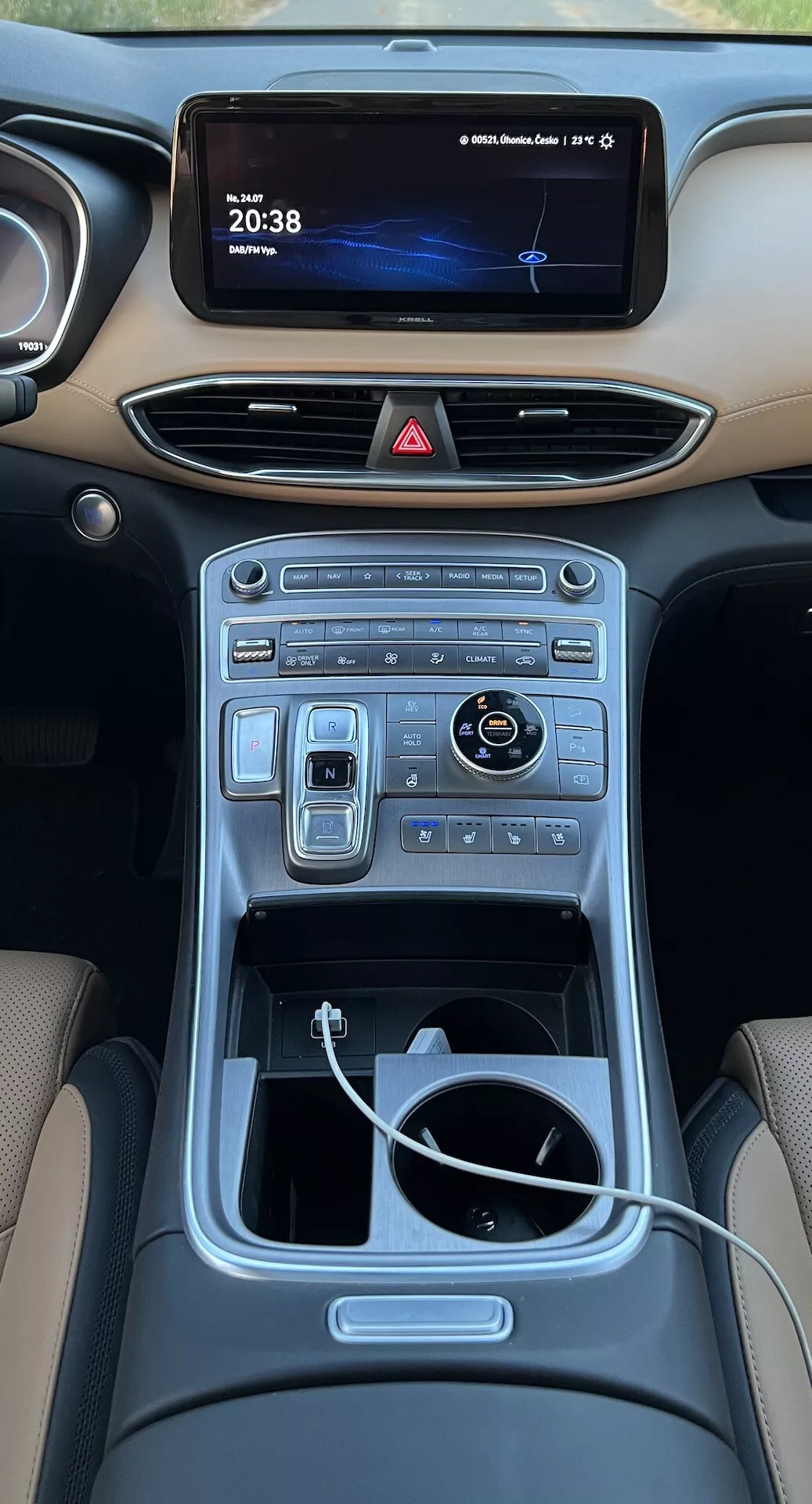 Vista desde arriba del interior de la consola central del Hyundai Santa Fe 2020 desde los asientos traseros. La imagen muestra un tablero moderno con una carretera nocturna en la pantalla de infoentretenimiento y un reloj digital que marca las '20:38.' Debajo, las rejillas de ventilación horizontales se integran perfectamente en el tablero. La consola central incluye una serie de controles táctiles y digitales para las funciones de clima y multimedia. Un dial giratorio brillante y los interruptores adyacentes sugieren navegación avanzada y modos de conducción. Más abajo, un área de almacenamiento abierta con un cable blanco visible indica conectividad de dispositivos. El túnel central, bordeado con una moldura metálica, mejora el diseño interior lujoso.