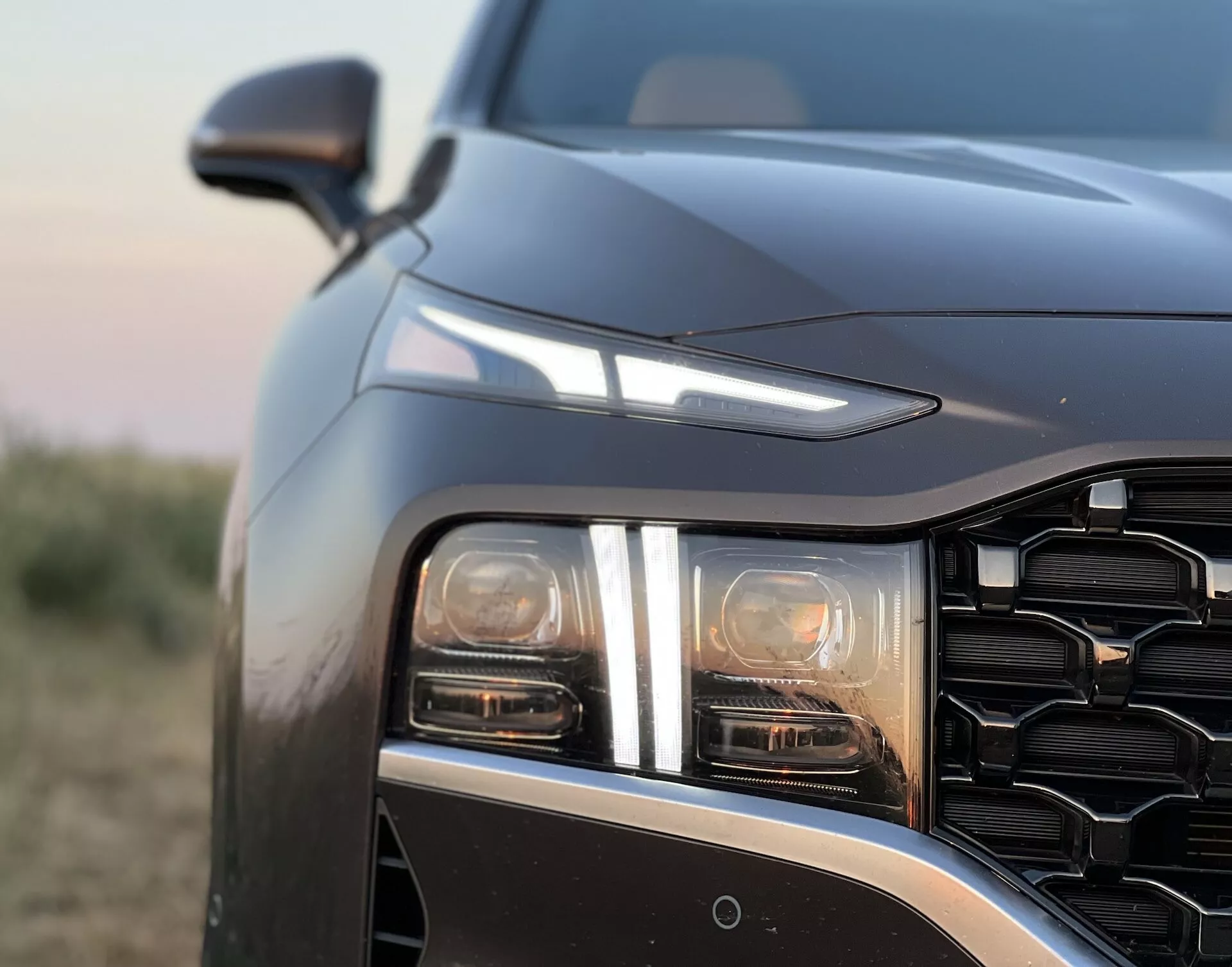 Vista detallada de cerca del Hyundai Santa Fe 2020, enfocándose en el cuarto delantero izquierdo desde un ángulo ligeramente frontal y bajo. La imagen enfatiza el grupo de faros LED o HID con al menos dos elementos de luz distintos, posiblemente incluyendo un haz principal y luz diurna o indicador de giro. La rejilla con patrón geométrico y la línea de capó que cae bruscamente sugieren un diseño contemporáneo y agresivo. El espejo lateral es visible más allá de la línea del capó, insinuando características tecnológicas avanzadas como un sensor o cámara en el parachoques delantero, posiblemente para asistencia de estacionamiento o prevención de colisiones. La fotografía tomada durante el amanecer o atardecer muestra iluminación de bajo contraste con tonos cálidos y una profundidad de campo superficial que destaca los detalles del diseño del vehículo contra un fondo suavemente desenfocado.
