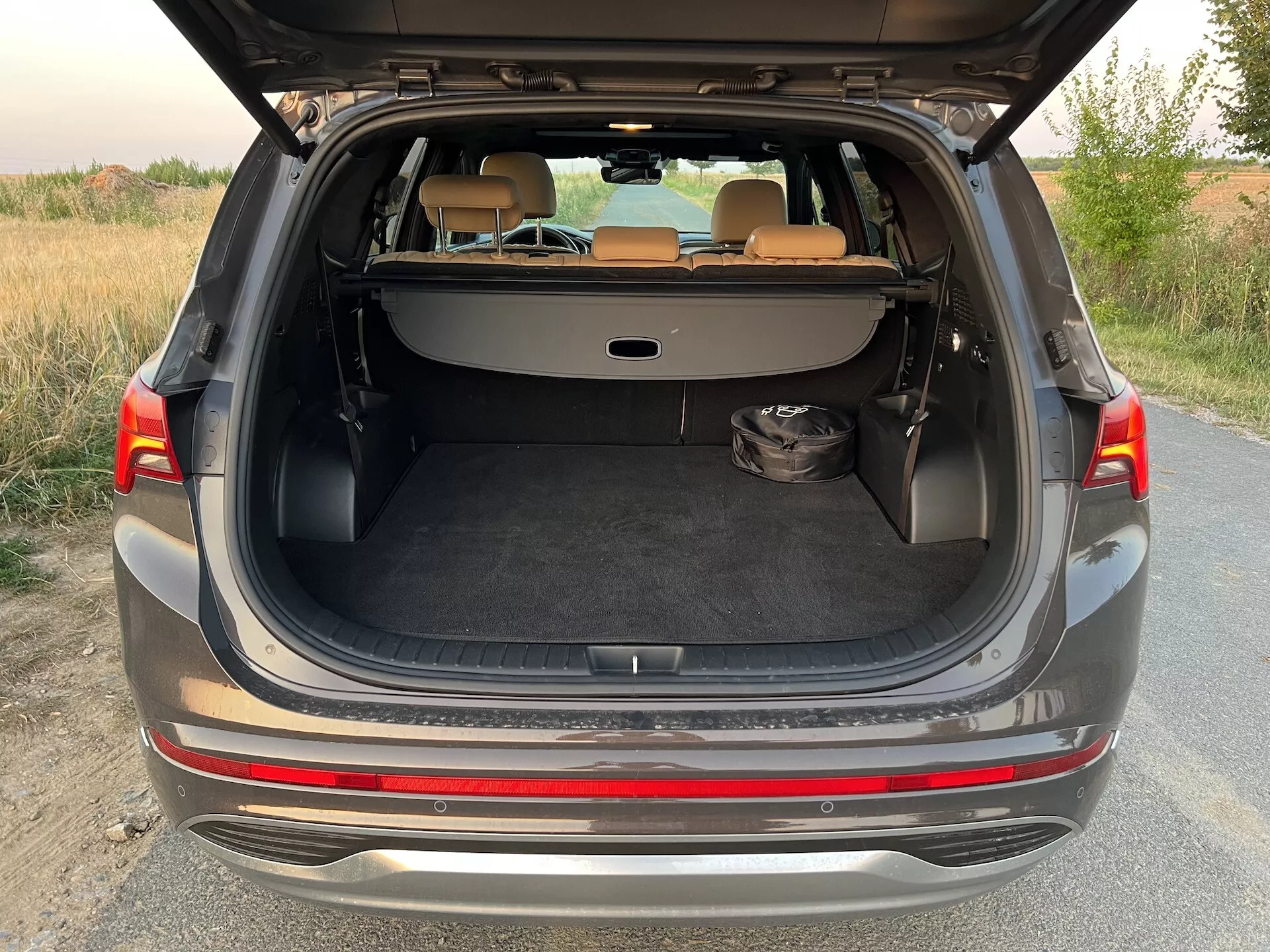 Sección trasera de un Hyundai Santa-Fe 2020 con el maletero tipo hatchback completamente abierto, mostrando el amplio espacio de carga. El vehículo, de color gris o plateado, está estacionado en un camino sin pavimentar rodeado de vegetación en un entorno rural o semi-rural durante una iluminación suave al amanecer o al anochecer. El diseño moderno presenta un parachoques bicolor, borde protector cromado de carga, piso de carga plano y puntos de sujeción para una cubierta de carga. Los asientos traseros están abatidos en una configuración de 60/40, proporcionando una superficie de carga extendida. La zona de carga está alfombrada y en el lado izquierdo se encuentra un objeto negro. El interior está en buenas condiciones y las luces traseras son parcialmente visibles.