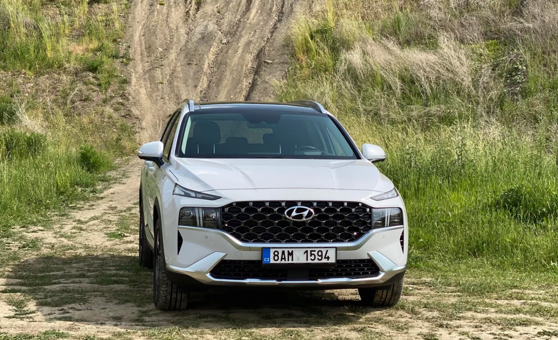 Vista frontal de un Hyundai Santa-Fe 2020 SUV posicionado en un camino de tierra con hierba y una colina en el fondo, mostrando la parrilla delantera con un patrón de malla geométrica, faros modernos y afilados con luces diurnas LED, y un parachoques delantero con tomas de aire. El capó presenta contornos acentuados y se ve una matrícula de estilo europeo. La imagen enfatiza el diseño y las características del vehículo en un entorno todoterreno.