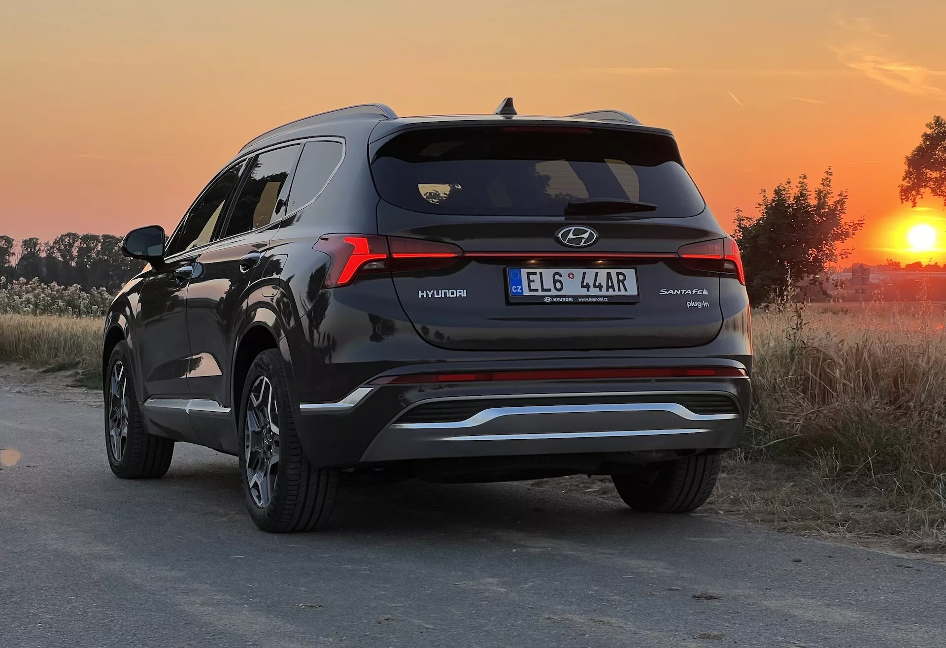 Vista trasera tres cuartos de un SUV Hyundai Santa Fe 2020, destacando el lado derecho y el perfil trasero con insignias visibles. La fotografía fue tomada durante la hora dorada, ofreciendo una luz de fondo cálida que acentúa el color de la pintura y los reflejos. El SUV está estacionado en un camino polvoriento o no pavimentado con un entorno rural. El ángulo bajo de la cámara, alrededor de la altura de las luces traseras, muestra el diseño de las luces traseras, la ventana trasera, los contornos de la tapa del maletero y las puntas de escape dobles. También son visibles detalles como una línea de acento metálico en el parachoques inferior, un alerón trasero y una antena de aleta de tiburón.