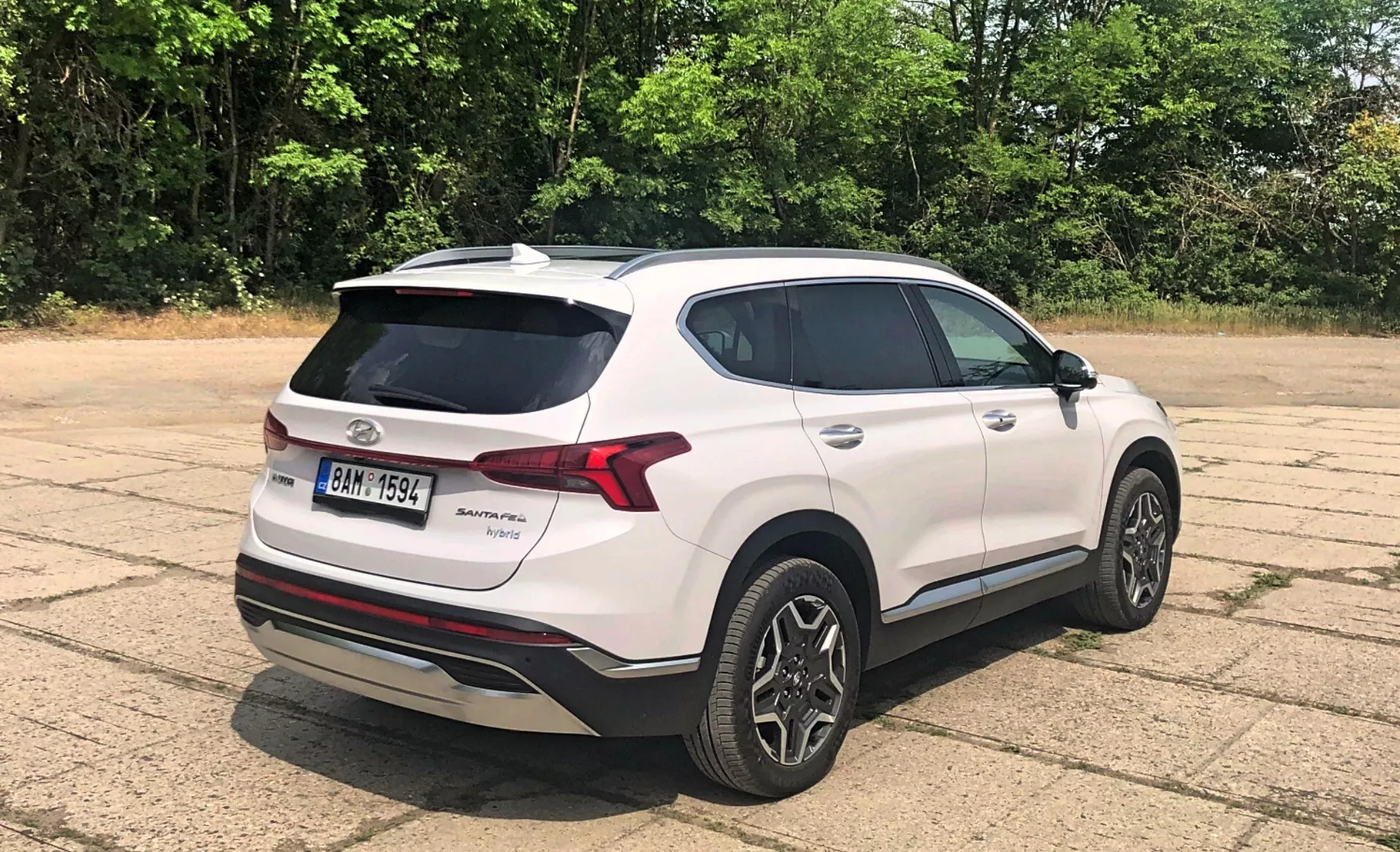 Vista trasera de tres cuartos del Hyundai Santa Fe 2020, un SUV crossover moderno blanco, estacionado al aire libre en un área pavimentada con árboles y vegetación de fondo bajo condiciones soleadas. Las características visibles incluyen luces traseras LED, alerón trasero, antena de aleta de tiburón, llantas de aleación bitono, vidrios polarizados, placa de marca y modelo en la puerta trasera, placa de protección plateada bajo el parachoques trasero, espejos laterales del color de la carrocería con luces intermitentes integradas y molduras cromadas alrededor de las ventanas.