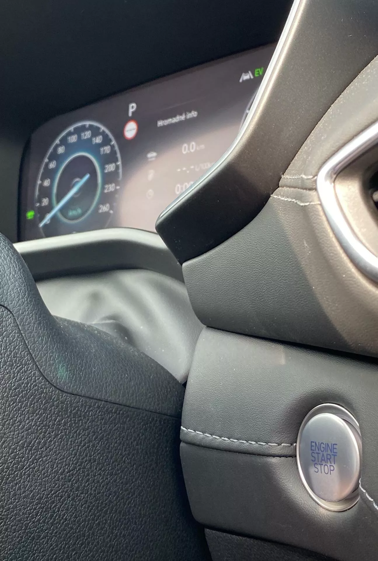 Vista interior de primer plano del Hyundai Santa Fe 2020, enfocándose en el lado izquierdo del volante de cuero y el panel de instrumentos digital. La foto, tomada desde el asiento del conductor e inclinada ligeramente hacia arriba, muestra el diseño moderno del volante, las costuras detalladas, el agarre contorneado y el encendido sin llave con un botón 'ENGINE START STOP'. El panel de instrumentos digital muestra el velocímetro indicando que el coche está detenido y otra información del vehículo, enfatizando un diseño premium y ergonómico.