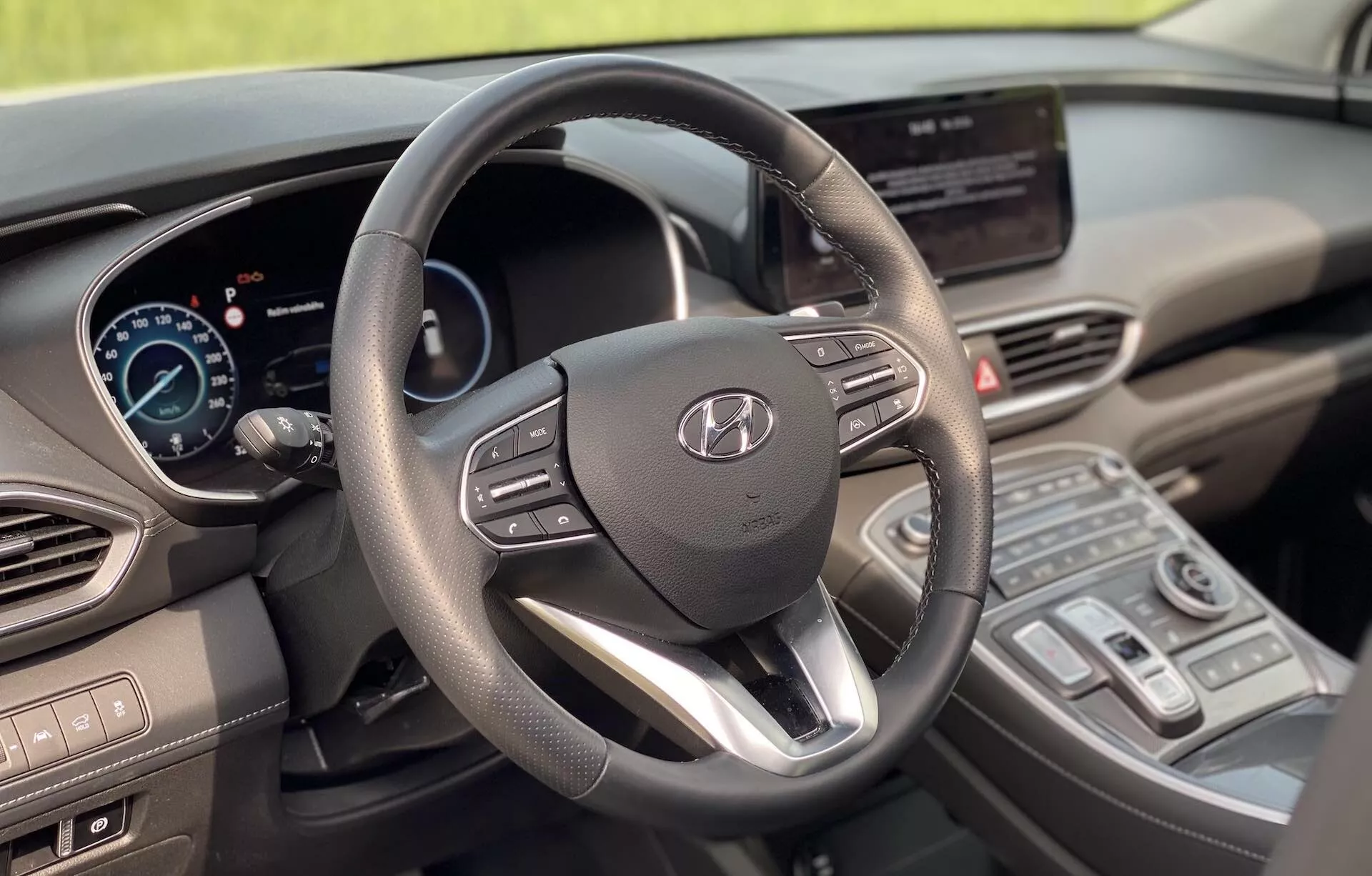 Foto del interior del Hyundai Santa Fe 2020 desde el asiento del conductor, destacando el volante moderno de tres radios con el logo de Hyundai y controles integrados, el panel de instrumentos digital, la pantalla central de infoentretenimiento táctil, la consola central con controles del clima y una palanca de cambios moderna. El diseño muestra un equilibrio entre funcionalidad y estética moderna con materiales suaves al tacto y acentos metálicos, indicando características tecnológicas avanzadas. La imagen muestra un vehículo limpio, bien mantenido y estacionario sin desenfoque de movimiento.