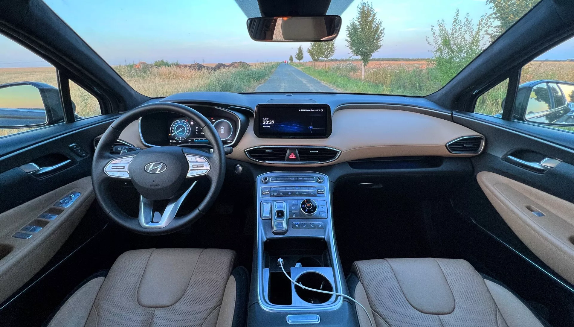 Vista de gran angular desde el asiento del conductor de un Hyundai Santa Fe 2020 que muestra los asientos de cuero beige, el tablero detallado con clúster de instrumentos digital, volante con la marca Hyundai y controles montados, sistema de infoentretenimiento con pantalla, y una consola central completamente equipada con palanca de cambios, portavasos, perillas de control y compartimento de almacenamiento.