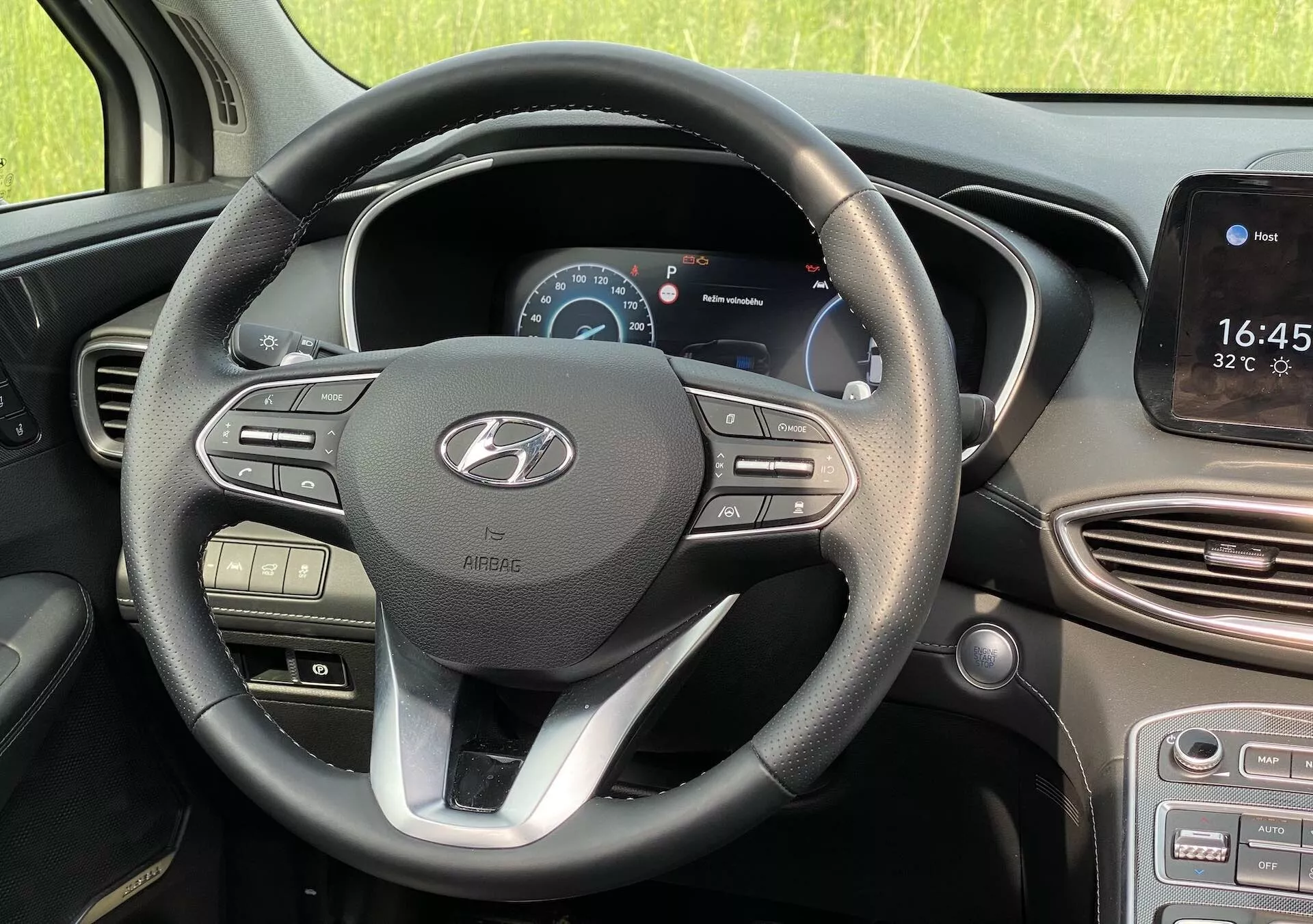 Vista interior de un Hyundai Santa Fe 2020 desde la perspectiva del asiento del conductor, mostrando el volante moderno de tres radios con acento plateado y el logo de Hyundai. El radio izquierdo tiene controles de volumen y medios, mientras que el radio derecho presenta botones de control de crucero y navegación de menús. Detrás del volante, un panel de instrumentos digital muestra la velocidad y el tacómetro. A la derecha, una pantalla táctil de infoentretenimiento muestra la hora y la temperatura, con posible navegación e integración de teléfonos inteligentes. Debajo hay rejillas de ventilación y botones de control de clima. El coche está estacionado a la luz del día con un entorno natural, no urbano visible a través de las ventanas.