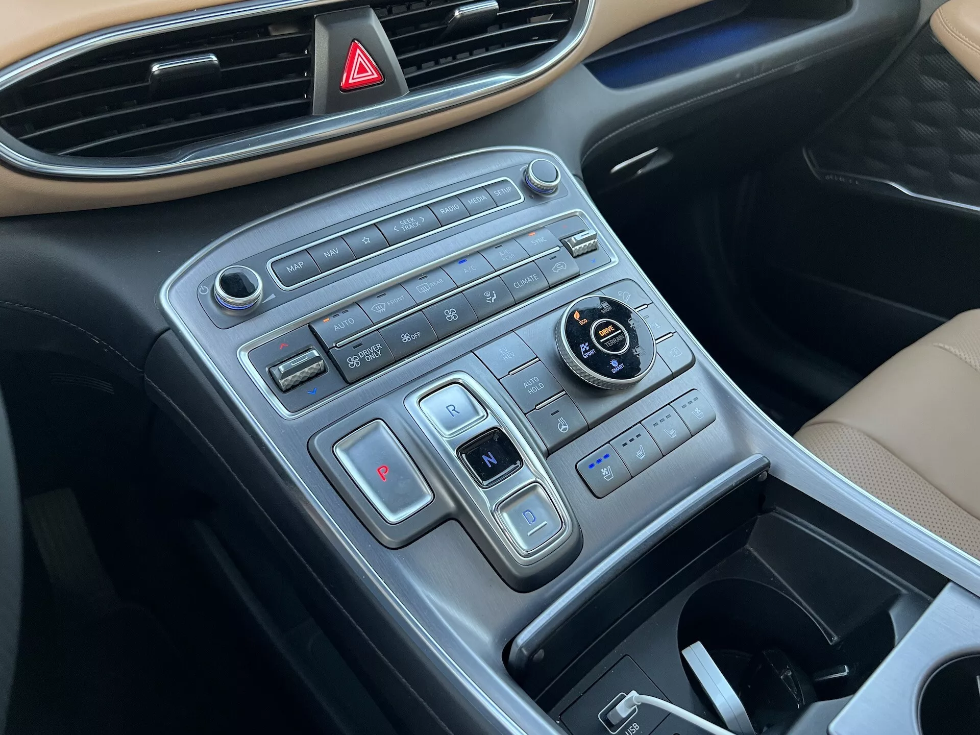 Vista de cerca de la consola central del Hyundai Santa Fe 2020 desde un ángulo del lado del pasajero, mostrando el tablero con el botón de luces de emergencia en la parte superior, controles de diseño moderno en filas horizontales que incluyen selector de modo de conducción, configuraciones de control de tracción, sistema de control climático de doble zona, botones de calefacción de asientos y un compartimiento de almacenamiento retráctil, con un acabado que imita el metal cepillado.