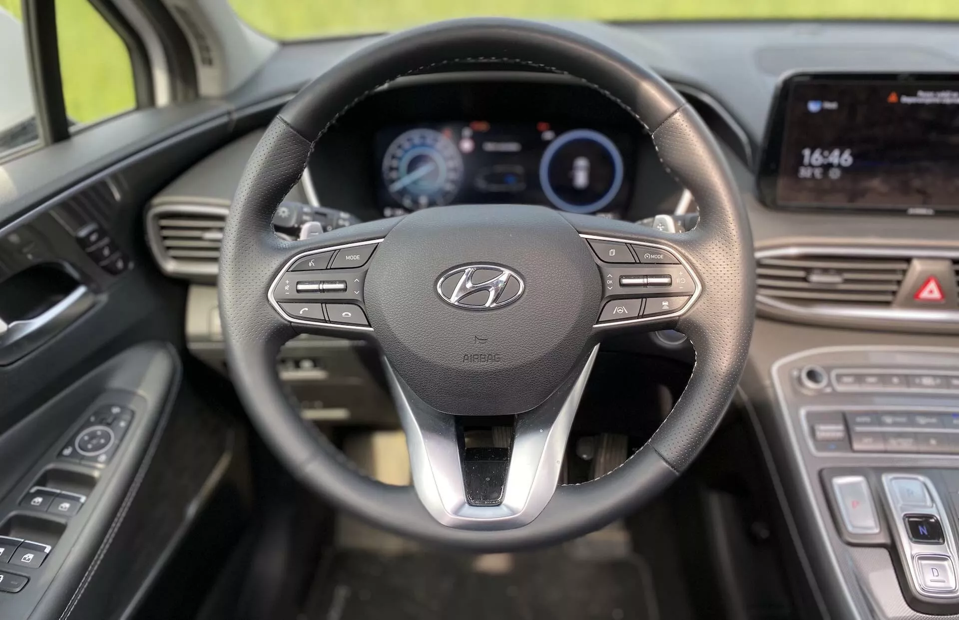 Perspectiva del conductor del interior del Hyundai Santa-Fe 2020 modelo híbrido, con un volante prominente que presenta el logotipo de Hyundai y botones de control para multimedia y conectividad. Detrás del volante hay un panel de instrumentos digital iluminado que muestra información clave como la velocidad y el consumo de combustible. A la derecha, la consola central incluye una pantalla táctil que muestra la hora (16:46) y probablemente es compatible con funciones de navegación y multimedia. Debajo de la pantalla táctil, se ven los controles de clima, lo que mejora el diseño moderno del vehículo. Los controles de las puertas para las ventanas eléctricas y los ajustes de los espejos laterales están ubicados a la izquierda del volante.