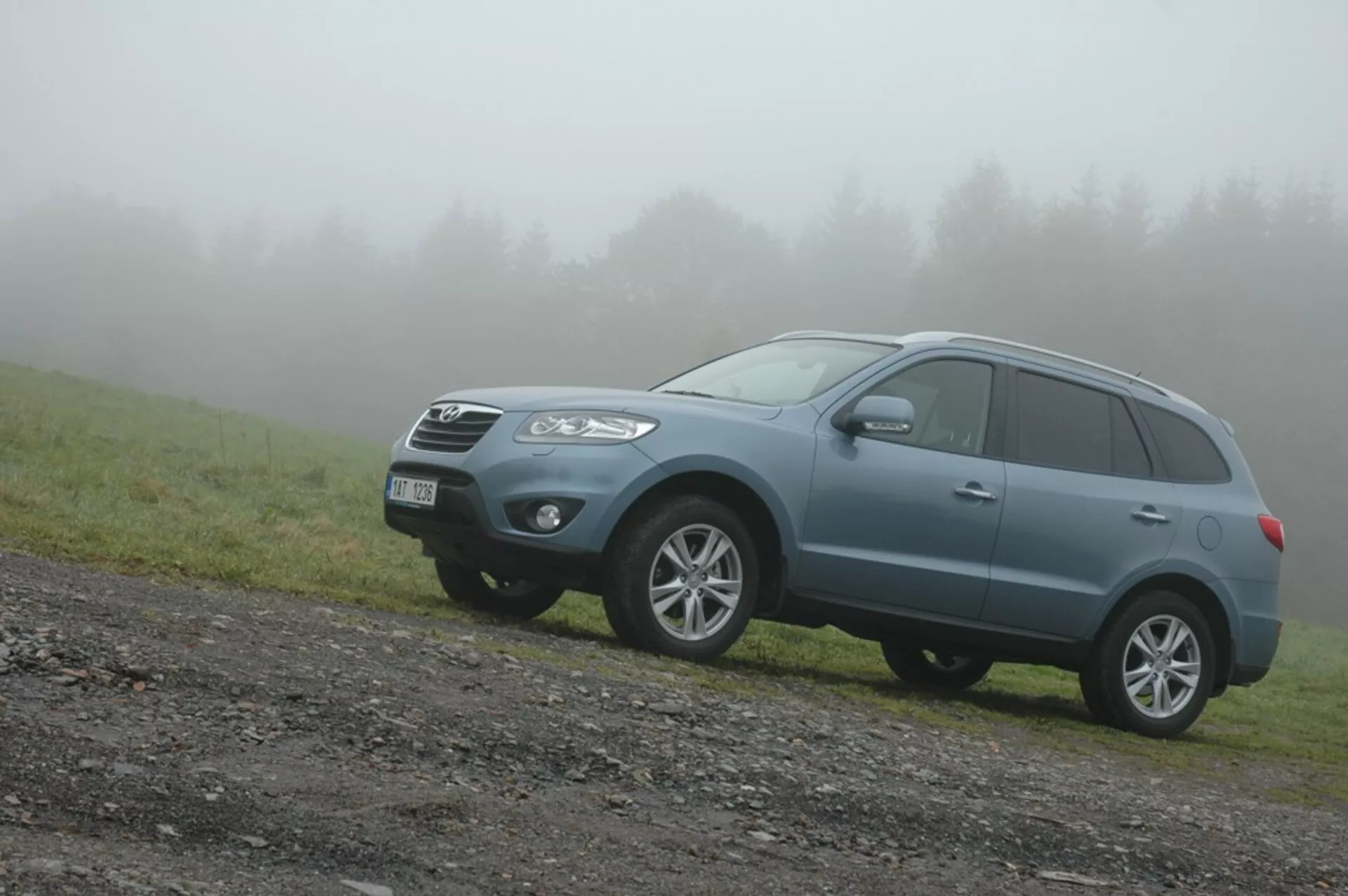 SUV crossover Hyundai Santa Fe azul de tamaño mediano del 2006 en un camino sin pavimentar con un parche de césped, vista frontal izquierda en ángulo en clima neblinoso destacando su diseño robusto, capacidades todoterreno y características de vehículo familiar moderno como parrilla frontal, faros angulares, luces de niebla, ruedas de aleación, barras de techo y señales de giro integradas.
