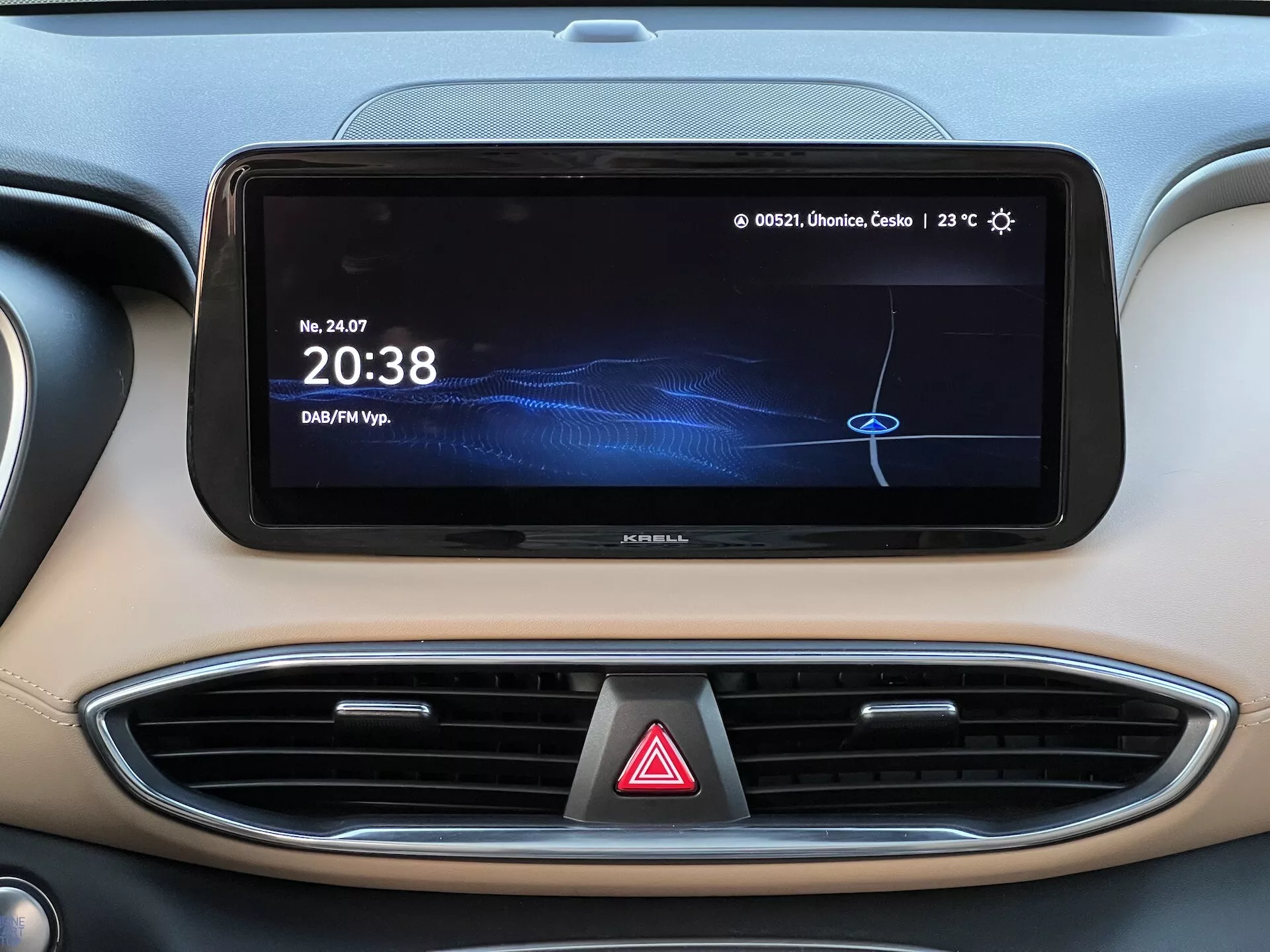 Primer plano del área central del tablero en un Hyundai Santa Fe 2020, mostrando la pantalla horizontal del sistema de infoentretenimiento con un reloj digital '20:38', fuente de medios etiquetada como 'DAF/MP3', lectura de temperatura '23°C' y texto parcial en polaco. También se observan dos salidas de aire con acento cromado y el botón de luces de emergencia debajo de la pantalla. El tablero presenta un esquema de color bicolor con una estética interior moderna y de alta calidad.
