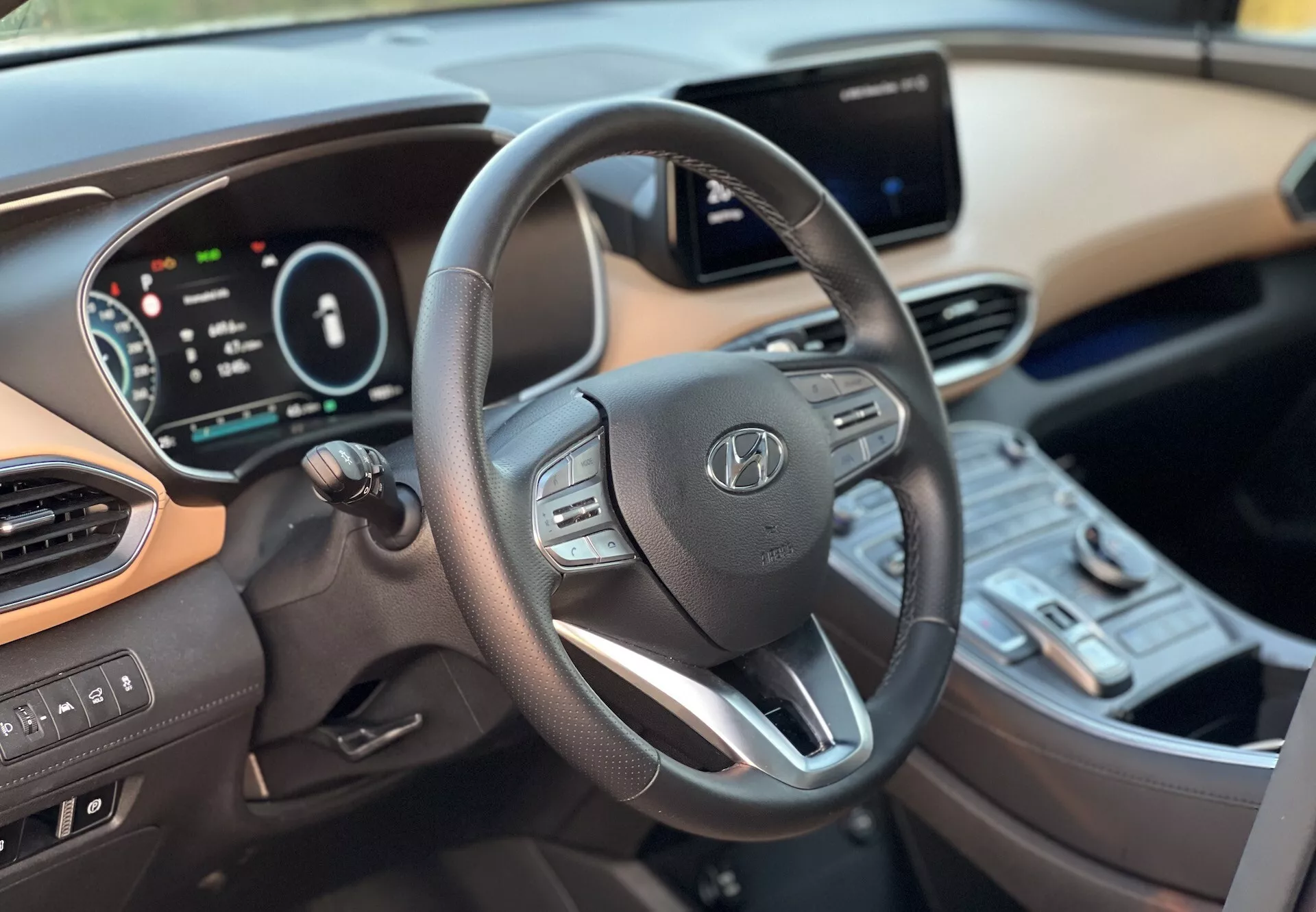 Vista interior del Hyundai Santa-Fe 2020 que muestra el lado del conductor desde un ángulo hacia la derecha del centro, enfocándose en el volante moderno de tres radios con controles integrados, el cuadro de instrumentos digital y un sistema de infoentretenimiento con pantalla táctil en la consola central. También se pueden ver los interruptores de control climático, el botón de arranque y el esquema de color dual del tablero y los interiores, ilustrando la tecnología avanzada y la comodidad del vehículo.