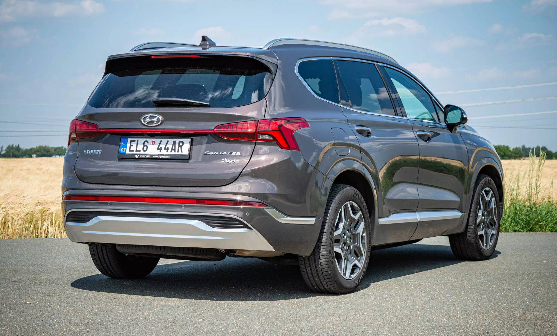 Vista trasera de tres cuartos de un Hyundai Santa Fe Hybrid 2020. La toma en ángulo bajo enfatiza la altura y robustez del SUV, con el coche posicionado en el lado derecho sugiriendo movimiento. Estacionado en una carretera pavimentada en un entorno rural, la parte trasera presenta luces traseras LED dispuestas verticalmente, un alerón trasero para estabilidad aerodinámica, tubos de escape dobles, alerón dividido en el techo y molduras reflectantes en el parachoques. El diseño insinúa sensores de estacionamiento traseros, cámara de visión trasera, ruedas de aleación de varios radios y una antena de aleta de tiburón para radio satelital o tecnología GPS.