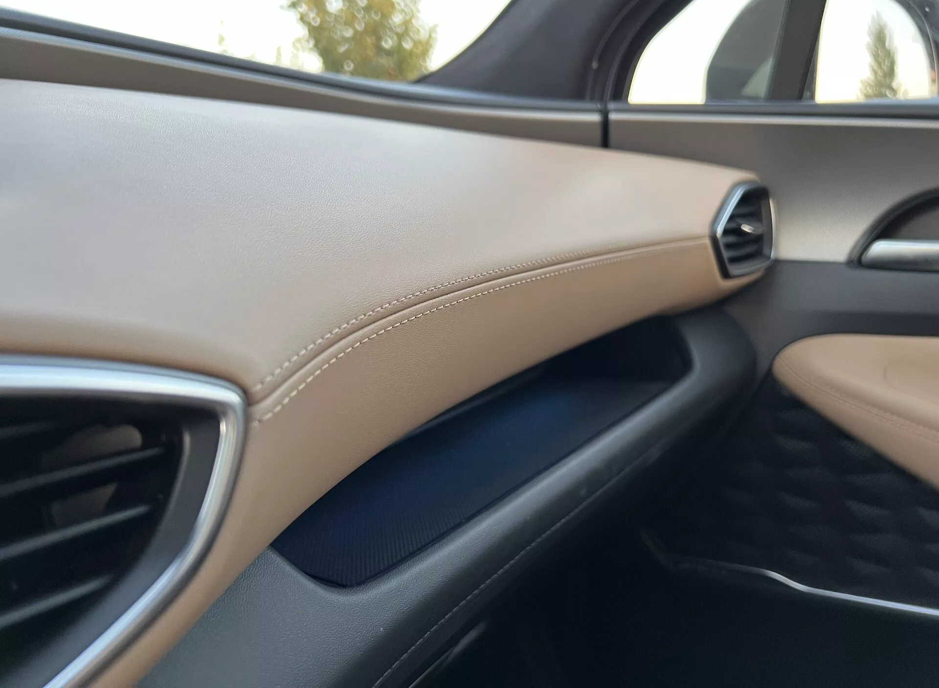 Detalle del interior del Hyundai Santa Fe 2020 que muestra el lado del pasajero del tablero. La imagen, tomada desde el asiento del conductor o del pasajero central, destaca una ventilación lateral derecha con borde metálico. Debajo del ventilador, se puede ver una superficie texturizada de color gris oscuro mientras que un panel beige con costura decorativa acentúa el acabado de lujo. El enfoque es suave, dando una impresión general del diseño interior, materiales y calidad de construcción. A través de la ventana se infiere que es de día, aunque los detalles exteriores no se ven claros.