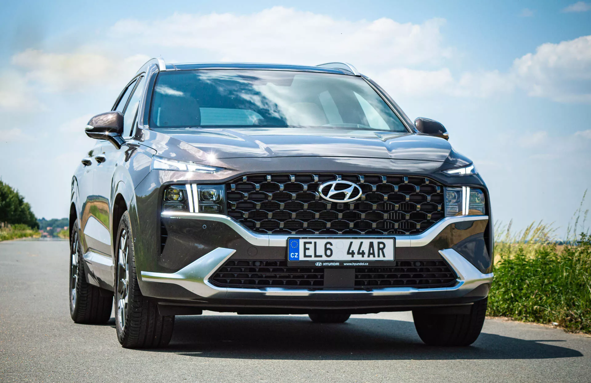 Vista de ángulo frontal de un SUV moderno, específicamente un Hyundai Santa Fe 2020, tomada desde un punto bajo cerca del lado del conductor. La cámara capta el audaz diseño frontal del vehículo, que incluye una pintura metálica negra o gris oscuro con un brillo reflectante, una rejilla de patrón en forma de panal con un emblema central de Hyundai, faros angulares y afilados con luces diurnas LED, iluminación adicional en el parachoques inferior como luces antiniebla, una gran entrada de aire y líneas esculpidas en el capó. El parabrisas es grande e inclinado, con espejos laterales parcialmente visibles y señales de giro integradas. La matrícula está montada en el centro de la parte inferior del parachoques.