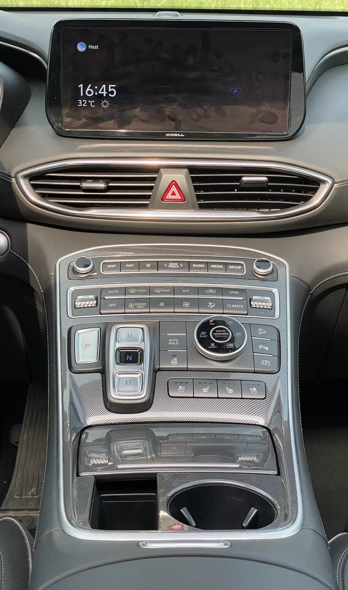 Imagen de la consola central del interior del Hyundai Santa Fe 2020, mostrando varios controles y pantallas en el tablero desde una perspectiva a nivel de los ojos. En la parte superior hay una pantalla táctil de infoentretenimiento orientada horizontalmente que muestra '16:45' y '32°C', con una superficie brillante que refleja el interior. Abajo hay ventilas de aire horizontales con acentos cromados y un botón rojo de luces de emergencia. La consola tiene botones y diales, incluido un sistema de control climático de doble zona, controles de temperatura de los asientos y más controles de funciones del vehículo como modos de conducción y sensores de estacionamiento. Hay asas a cada lado, enfatizando deportividad o capacidad todoterreno.