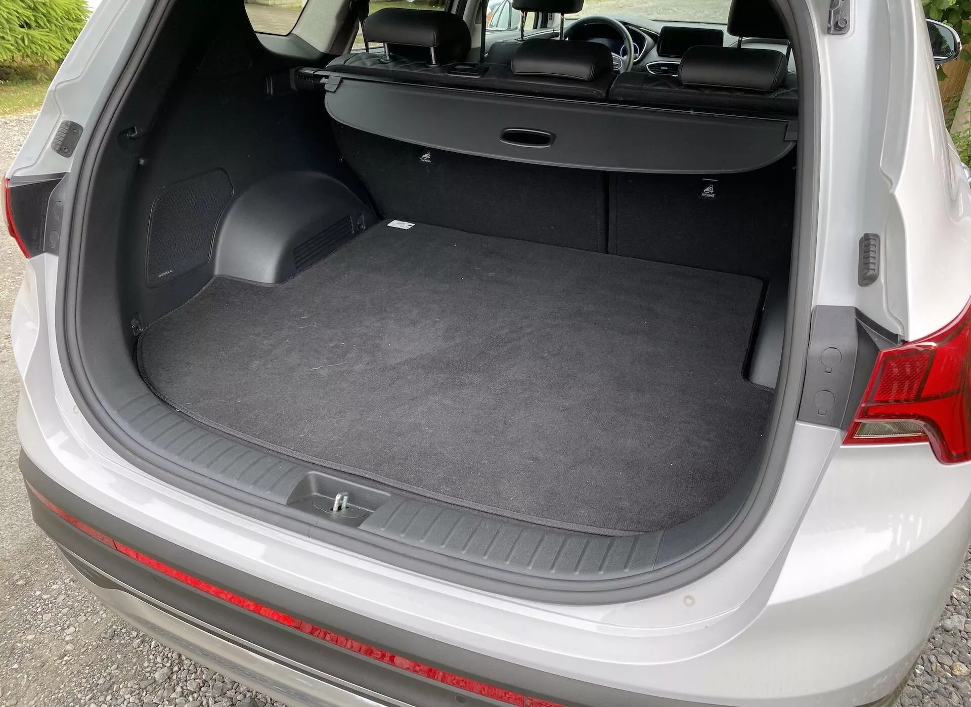 Toma en ángulo alto del área de carga de un Hyundai Santa-Fe 2020, vista desde la parte trasera con la escotilla abierta. La imagen muestra un suelo de carga plano, superficie alfombrada y paredes laterales con pequeños compartimentos de almacenamiento y ganchos para accesorios. Los asientos traseros están plegados en posición vertical y la cubierta retráctil del maletero está empujada hacia atrás, destacando el interior versátil del vehículo.