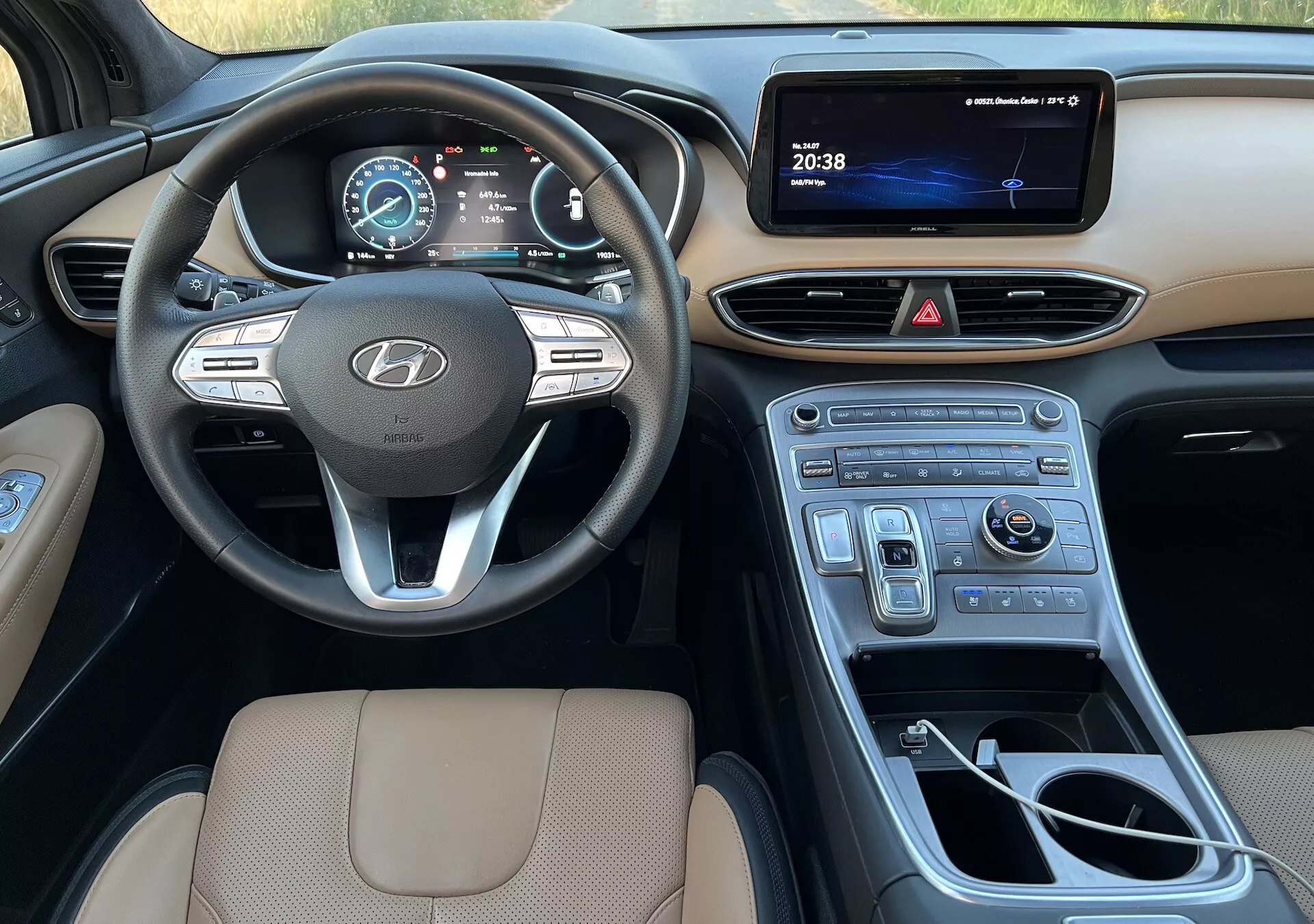 Esta imagen muestra el interior de un Hyundai Santa-Fe 2020, enfocándose en el lado del conductor. Ofrece una vista completa desde el asiento del conductor hasta la consola central, y se extiende al lado del pasajero, destacando varios elementos clave. El volante con el logotipo de Hyundai, con controles para audio, control de crucero y sistema de manos libres, se muestra prominentemente. Detrás de él, un cuadro de instrumentos digital muestra el velocímetro y la información del vehículo. La consola central incluye una pantalla de infoentretenimiento, botones de control para las luces de emergencia y el control del clima, y un adorno brillante de tipo madera. También se ve la palanca de cambios para la transmisión automática, un dial de control para navegación o multimedia, y portavasos. La parte superior del asiento del conductor, tapizada en un material color beige con costuras, enfatiza la comodidad y la calidad del diseño. La imagen captura el diseño moderno, fácil de usar, y las características tecnológicas del interior del vehículo durante el día con iluminación natural.