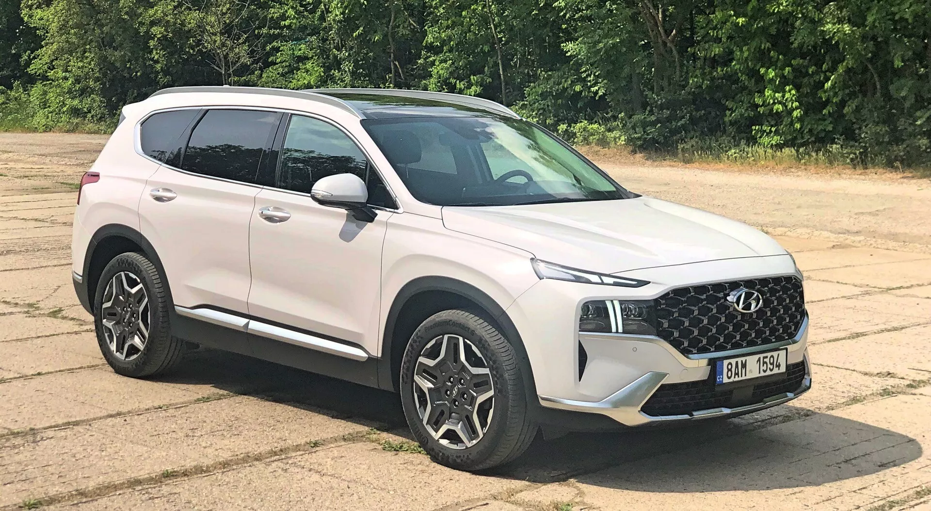 SUV moderno Hyundai Santa Fe 2020 estacionado en superficie de asfalto visto desde la parte frontal de tres cuartos, mostrando diseño de parrilla, faros elegantes, ruedas de aleación de múltiples radios, espejos laterales del color del cuerpo con señales integradas, líneas de carácter, perfil aerodinámico, pintura blanca brillante, ventanas ligeramente tintadas e iluminación natural y uniforme con vegetación en el fondo.