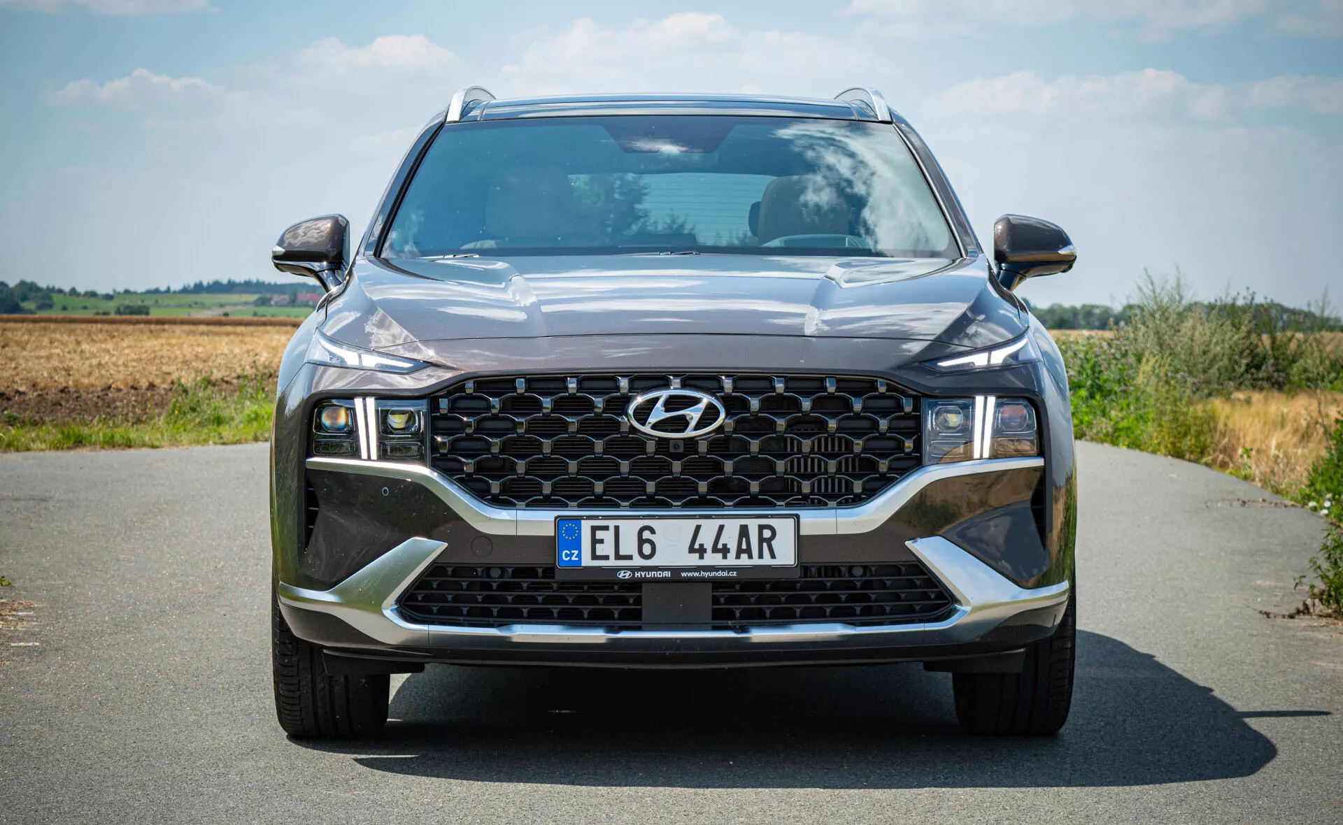 Hyundai Santa Fe 2020 - Vista frontal de un SUV moderno en una carretera asfaltada con un paisaje natural de fondo. Esta imagen en ángulo bajo resalta la imponente presencia del SUV, destacando detalles como la parrilla frontal hexagonal con patrón en panal, faros LED angulares, acentos contrastantes en el paragolpes delantero y elementos de iluminación adicionales, incluyendo luces antiniebla.
