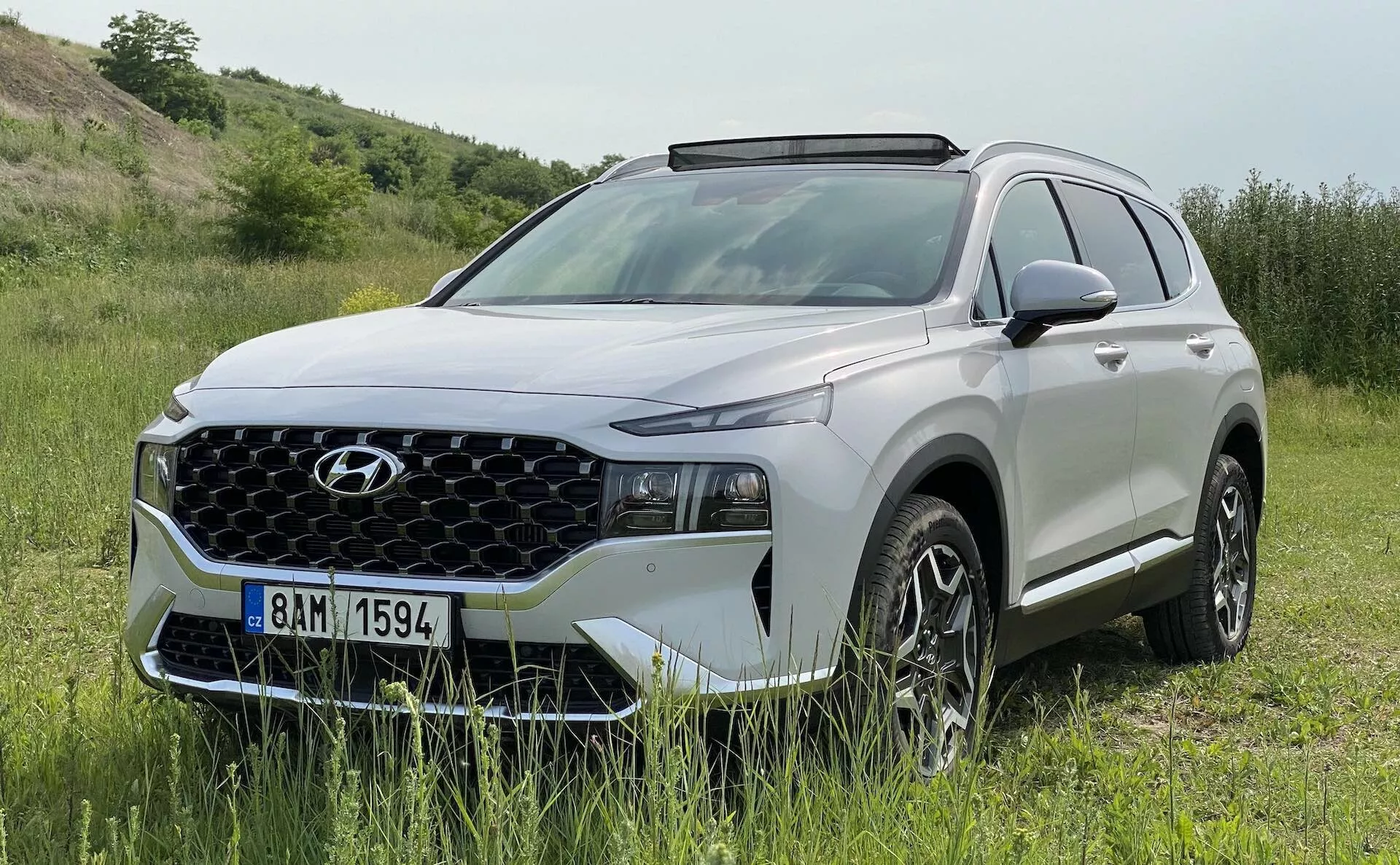 Un SUV Hyundai Santa Fe 2020 moderno en un entorno natural al aire libre. El vehículo está estacionado en un área de pasto con una ligera inclinación, sugiriendo capacidades todoterreno. Imagen diurna brillante que muestra el perfil delantero y lateral con características como una prominente parrilla geométrica, faros LED estrechos, luces diurnas, llantas de aleación con diseño de múltiples radios, espejos laterales del color de la carrocería con señales de giro integradas, rieles de techo y revestimiento plástico alrededor de los arcos de las ruedas. La imagen captura el diseño y potencial aventurero del SUV, destacando su utilidad y aspecto premium.