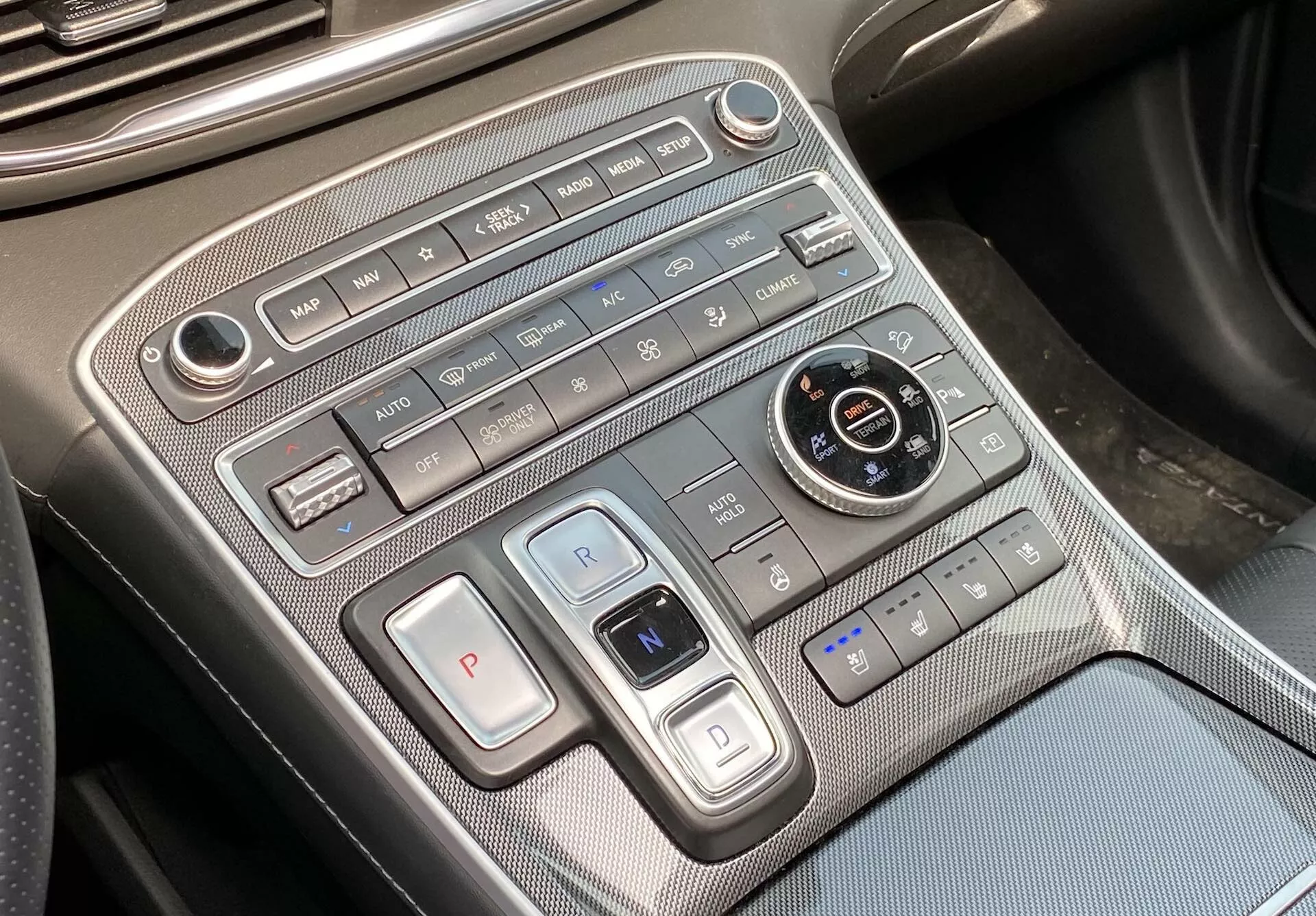 Esta imagen muestra una vista de primer plano de la consola central del Hyundai Santa Fe 2020, enfocándose en el área de control de clima y transmisión. La toma, ligeramente en ángulo desde la perspectiva del conductor o pasajero, revela el panel de control de clima dispuesto horizontalmente en la parte superior, que incluye controles de velocidad del ventilador, botones de flujo de aire, control de clima automático, descongelación y recirculación de aire. Debajo, hay botones de selección de marcha con un gran 'P' para parqueo y una palanca como interruptor para las marchas 'R' y 'N', lo que indica el uso de un sistema electrónico o de botones para la selección de marchas. Un dial redondo para los modos de conducción, rodeado de botones de auto-hold de freno, asistente de estacionamiento, y sistema de arranque/parada automático está presente. Los controles de los asientos para el calentamiento o ventilación son visibles en la parte inferior derecha. La consola cuenta con materiales premium con acentos metálicos y un patrón similar a la fibra de carbono. Un puerto USB en la parte inferior izquierda ofrece opciones de conectividad para dispositivos.