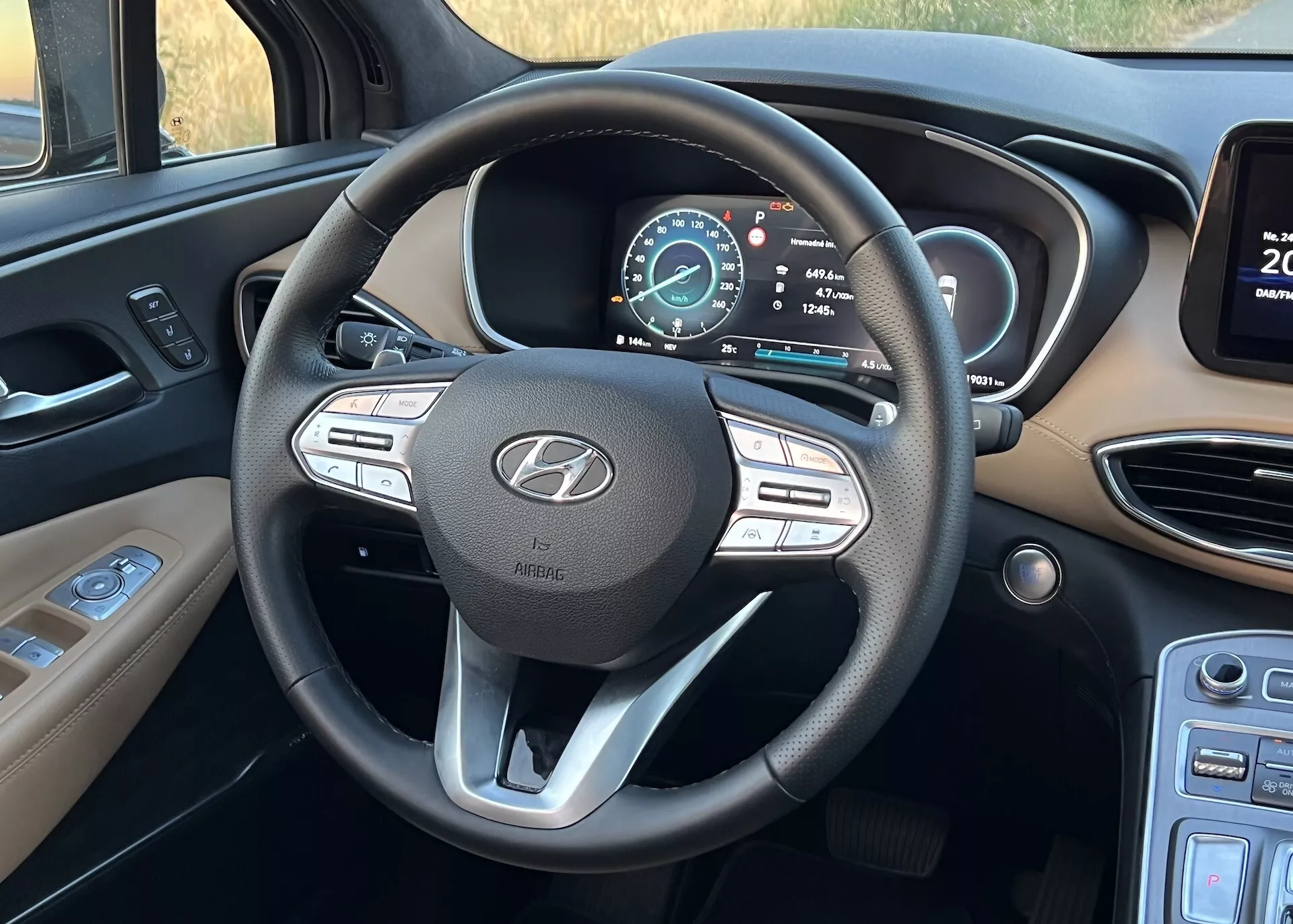 Imagen del interior del Hyundai Santa Fe 2020 centrada en el volante multifuncional de tres radios y el cuadro de instrumentos digital. Foto tomada desde la perspectiva del asiento del conductor, mostrando los botones de control de medios y crucero en el volante, el emblema de Hyundai con el texto 'AIRBAG' y un tablero digital multifuncional que indica que el vehículo está en modo de aparcamiento con una advertencia de puerta abierta. A la izquierda están los controles probables de las luces, y a la derecha, el borde de una pantalla de infoentretenimiento junto a las rejillas de ventilación.
