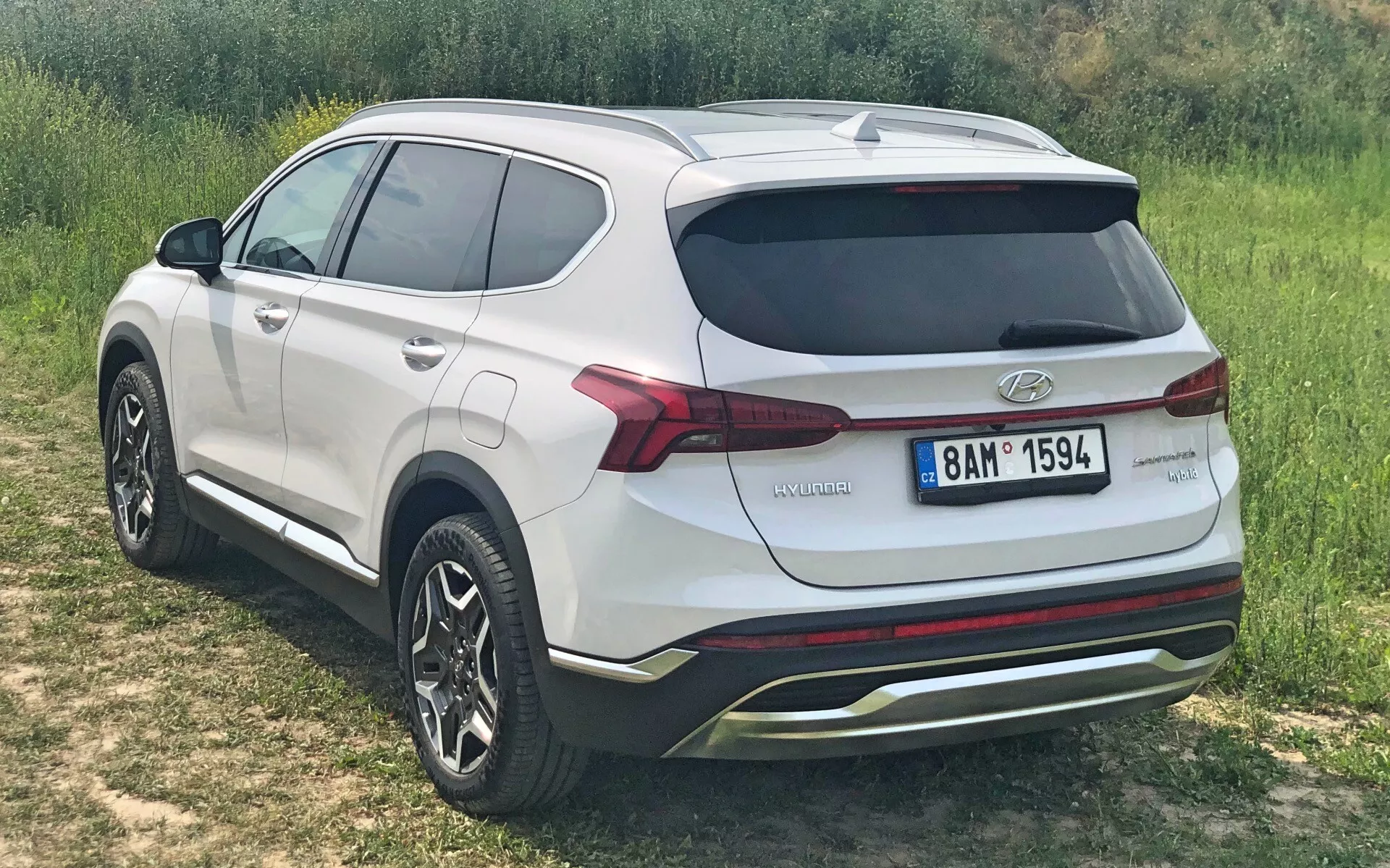 Una toma elevada de tres cuartos traseros de un Hyundai Santa Fe Hybrid 2020, que muestra la elegante línea del techo y el diseño del portón trasero. El SUV, estacionado en un terreno con césped, destaca su exterior moderno con un spoiler en el techo visible, líneas de contorno curvas y paragolpes traseros esculpidos. El vehículo cuenta con llantas de aleación de múltiples radios, luces traseras LED y una insignia que indica su tecnología híbrida. Pintado en un color blanco sólido con una apariencia limpia y pulida, el automóvil también incluye un parachoques trasero con un elemento tipo difusor, líneas de desempañador en la ventana trasera y ninguna modificación visible, lo que sugiere una condición de fábrica.