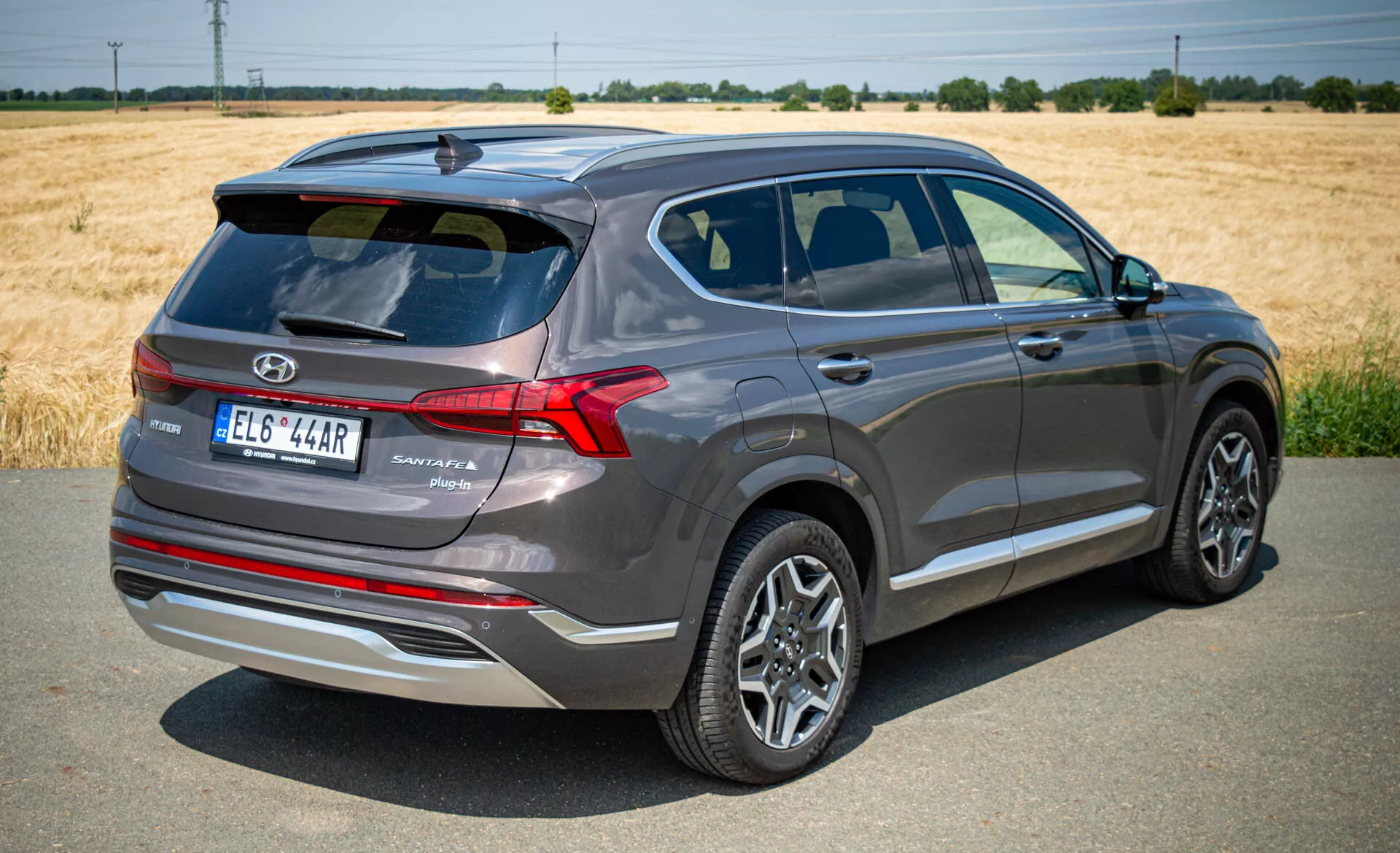 La imagen muestra una vista trasera de tres cuartos de un SUV moderno Hyundai Santa Fe 2020, capturada a plena luz del día bajo un cielo despejado. El coche está ligeramente inclinado a la derecha, revelando características clave como el portón trasero con el logotipo de la marca, el nombre del modelo y el nivel de acabado, luces traseras LED de diseño afilado, llantas de aleación con diseño multiradio, espejos laterales con luces intermitentes integradas y líneas de carácter. Las ventanas están teñidas con un borde cromado y la línea del techo barrida añade un atractivo deportivo. Colocado sobre una superficie pavimentada con un campo y árboles en el fondo, el coche metálico gris oscuro parece nuevo o bien mantenido.