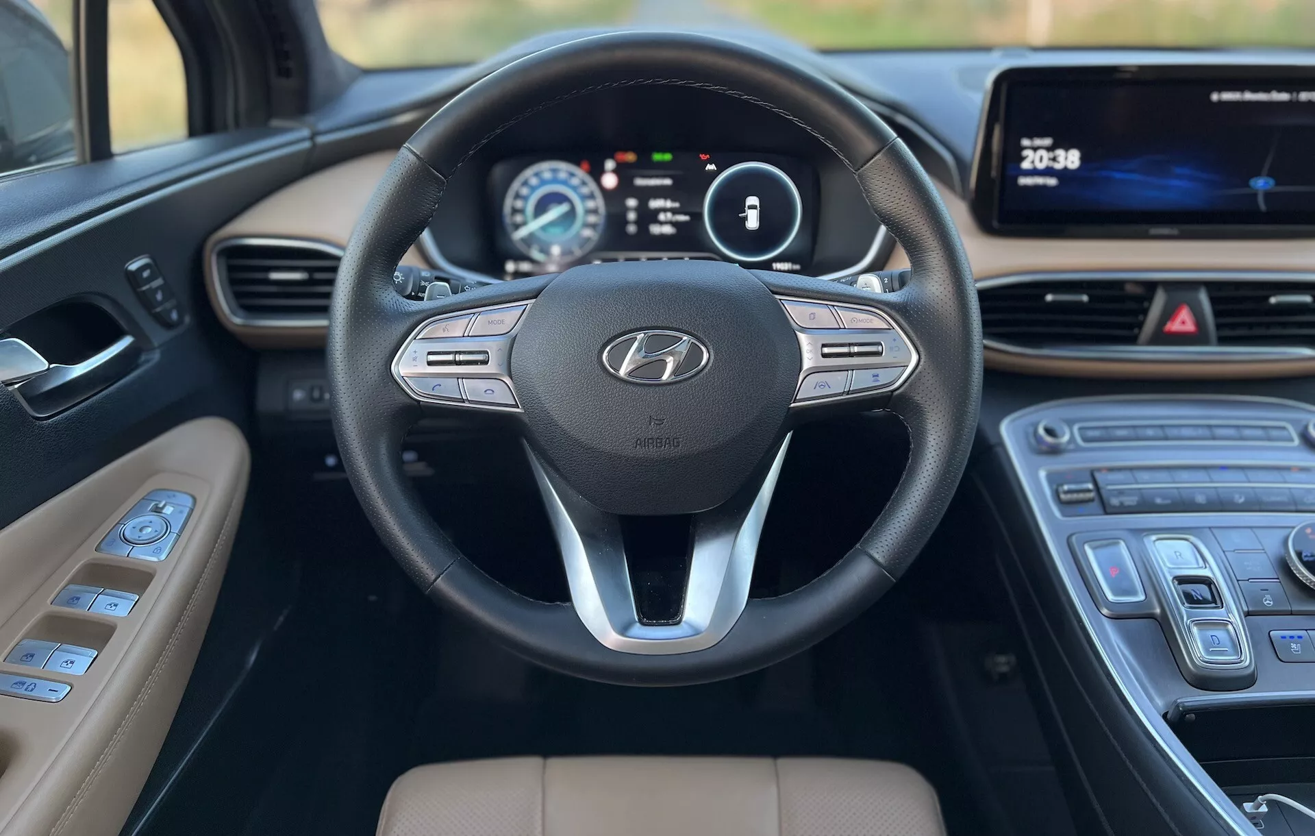 Vista de cerca desde los asientos traseros de un Hyundai Santa-Fe 2020, enfocándose en el tablero delantero y el volante. El volante presenta un diseño de tres radios con el emblema de Hyundai y botones de control para audio, comandos de voz y control de crucero. Detrás del volante, un clúster de instrumentos digital muestra la velocidad y las RPM en una pantalla colorida. La consola central incluye una pantalla táctil multimedia y un control de clima elegante con botones táctiles. La palanca de cambios tiene un diseño moderno con acentos plateados, y la tapicería marrón claro contrasta con el tablero y el volante oscuros, mostrando un interior enfocado en la tecnología y la comodidad.