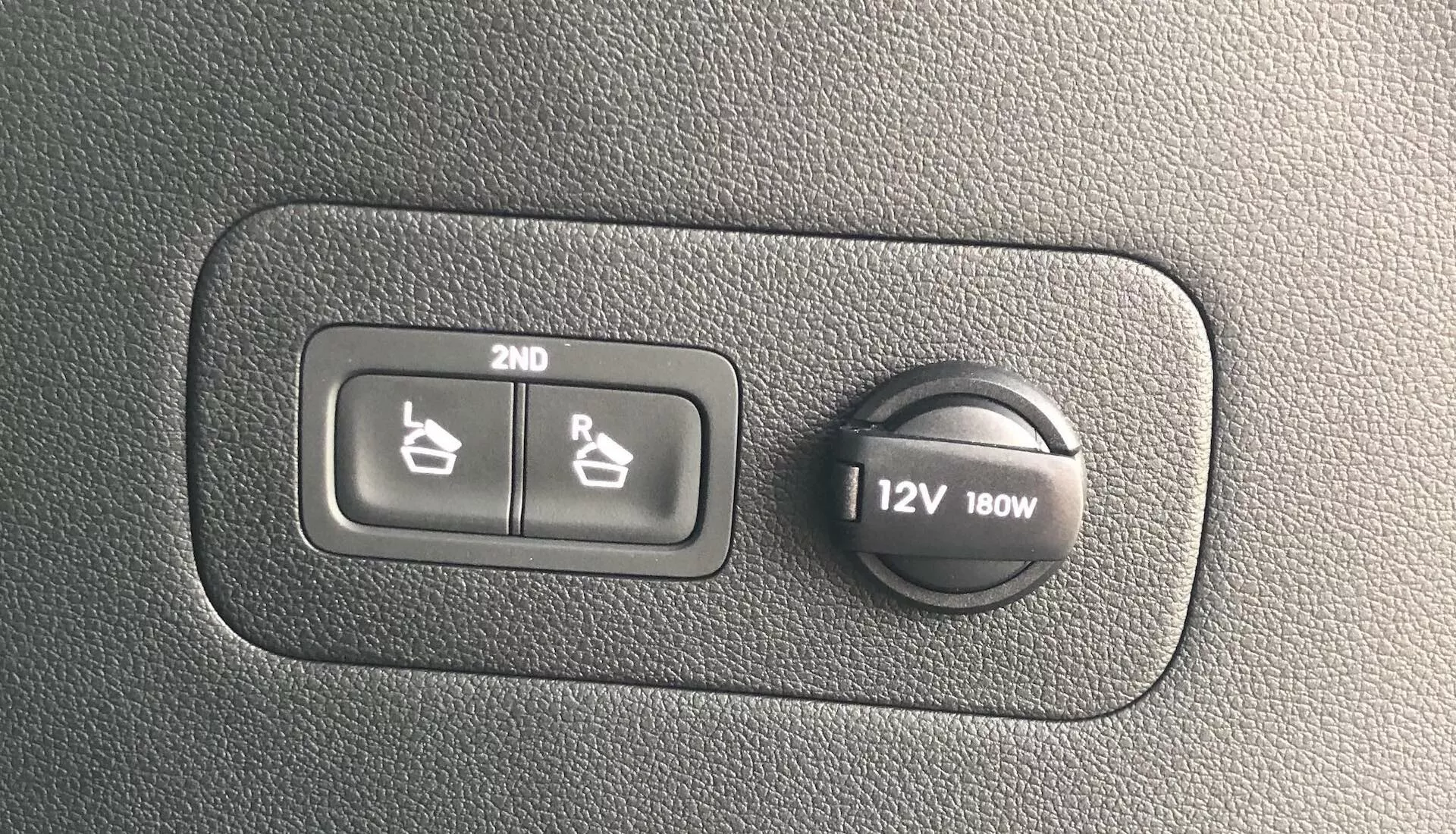 Primer plano de una sección del tablero del Hyundai Santa Fe 2020 que muestra los controles del calentador de asientos para la segunda fila y una toma de corriente de 12V 180W en una superficie de plástico texturizado.