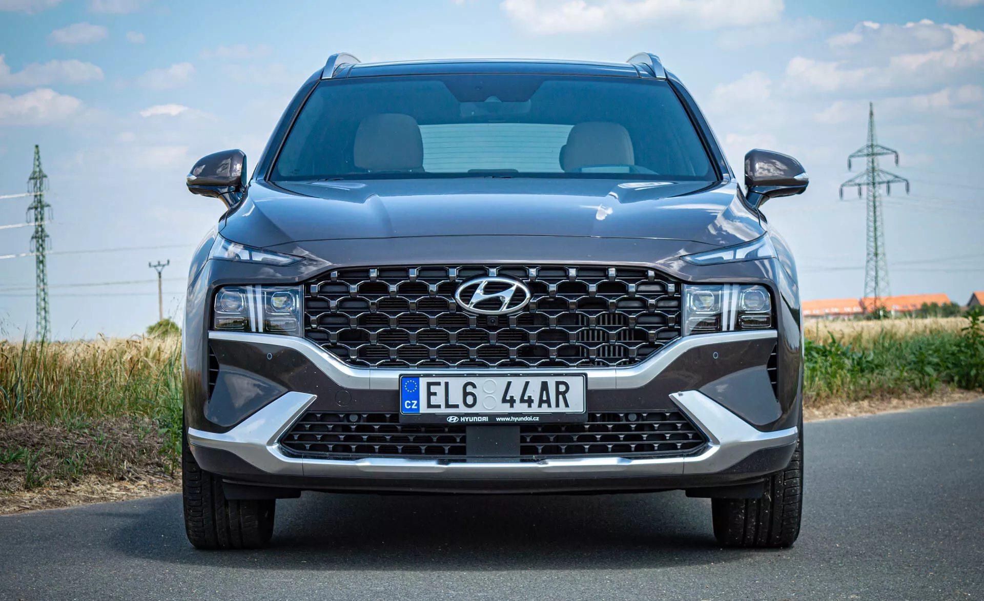 Vista frontal de un moderno SUV Hyundai Santa Fe 2020 tomada durante el día. La imagen muestra una gran parrilla en cascada con el logotipo central de Hyundai, matrícula de estilo europeo, elegantes faros LED angulares, amplia entrada de aire central y acentos plateados en el parachoques inferior. Son visibles las líneas esculpidas del capó, el parabrisas parcial y los espejos laterales con señales de giro integradas. La imagen enfatiza el tamaño del coche, detallando su diseño moderno y características tecnológicas.