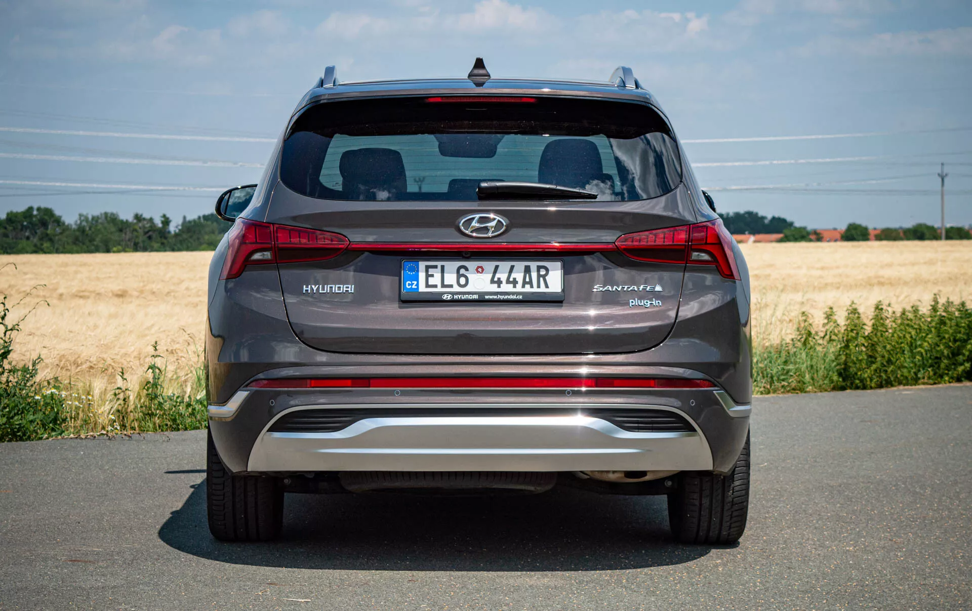 Vista trasera de un moderno SUV Hyundai Santa Fe 2020 plateado-gris estacionado en una carretera pavimentada, que muestra el logotipo de Hyundai, el nombre del modelo Santa Fe, matrícula de estilo europeo, luces traseras, parachoques trasero metálico reflectante con sensores de estacionamiento integrados, puntas de escape dobles, parabrisas trasero con alerón de marca Hyundai y tercera luz de freno. Clima despejado con un campo en el fondo.