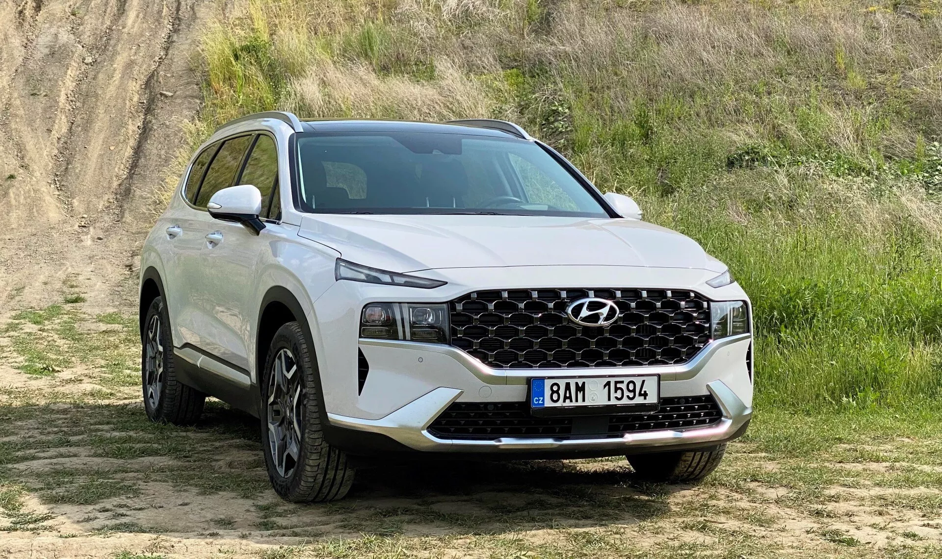 La imagen muestra un SUV Hyundai Santa Fe 2020 blanco estacionado sobre una superficie sin pavimentar con césped corto y un fondo elevado de tierra, sugiriendo un entorno de todoterreno ligero. Capturada desde una vista de tres cuartos delantera, la imagen detalla claramente la fascia frontal del vehículo, incluyendo una parrilla geométrica prominente que abarca casi todo el ancho, centrada con el logo de Hyundai y flanqueada por faros LED elegantes. El parachoques delantero inferior alberga iluminación adicional o sensores. Desde el perfil lateral hasta la puerta trasera del pasajero, grandes llantas de aleación llenan los arcos de las ruedas, y la carrocería superior presenta molduras que sugieren robustez. La silueta del SUV muestra una línea de techo descendente indicativa de diseños deportivos modernos. La escena está iluminada de manera natural, destacando las características del SUV sin sombras duras.